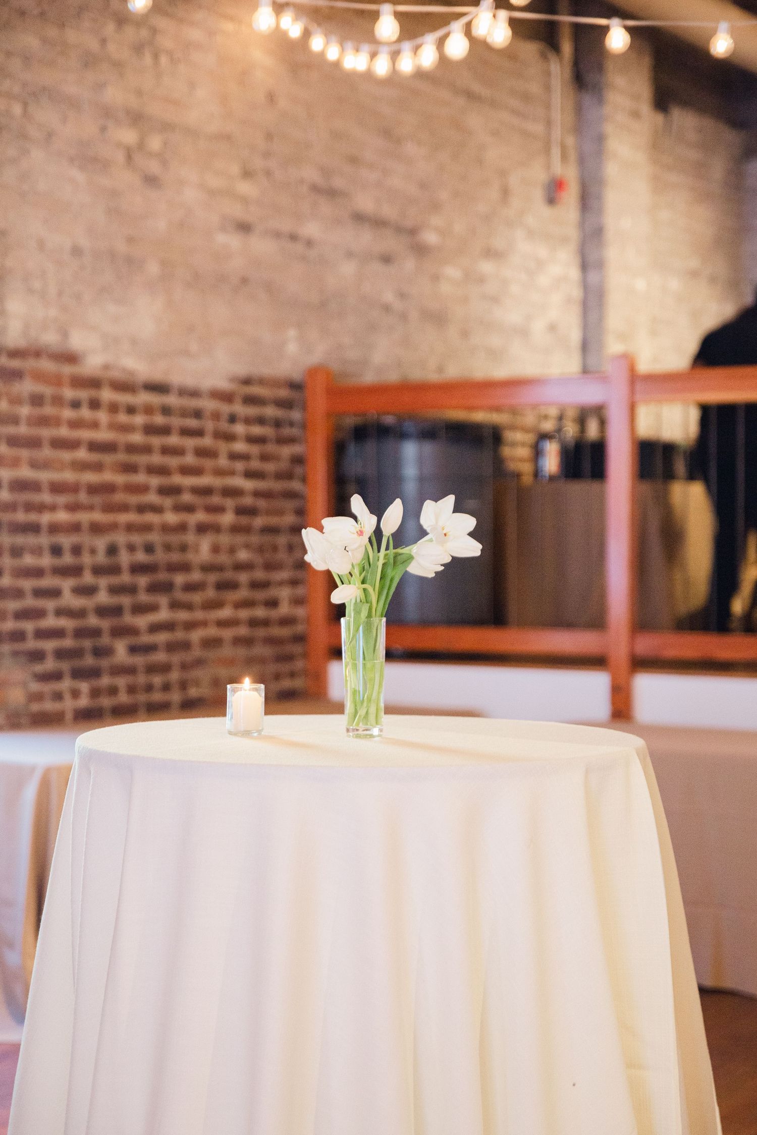 Cocktail table with white linen and tulip centerpiece under string lights in brick-walled venue.
