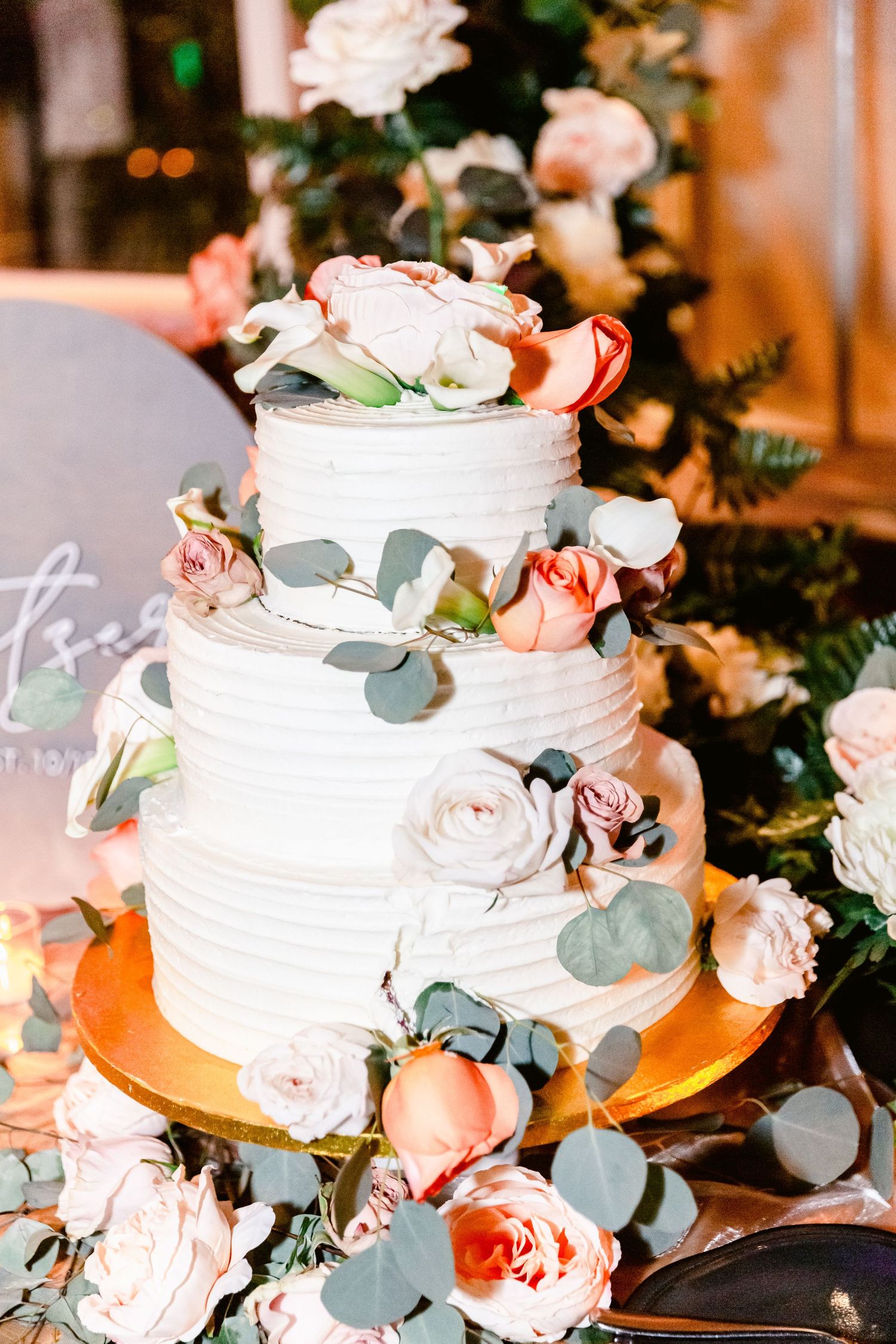 White tiered wedding cake decorated with blush roses and eucalyptus leaves on a rustic wooden table.