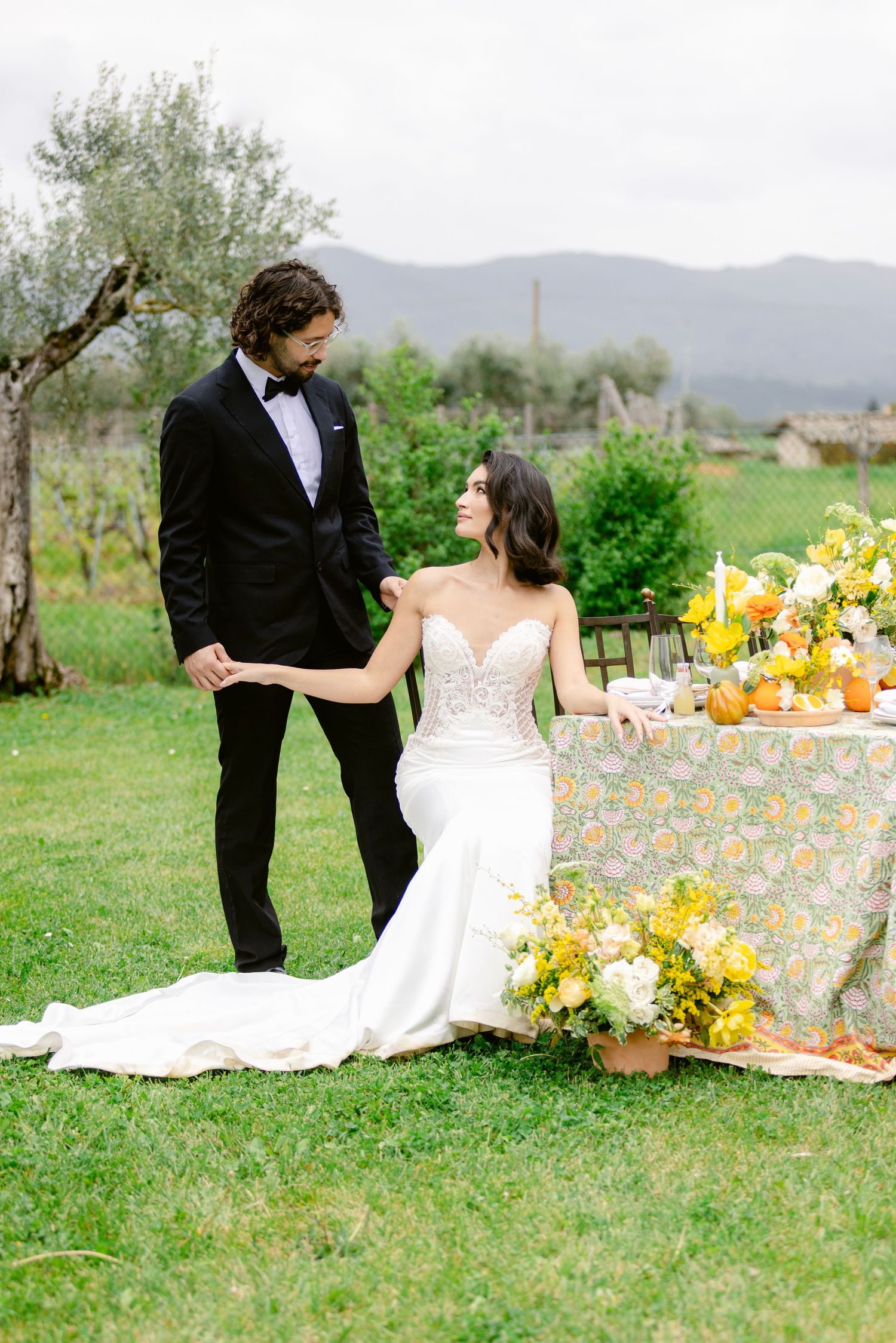 Couple in wedding attire having a romantic picnic outdoors with yellow floral decor and mountain views.