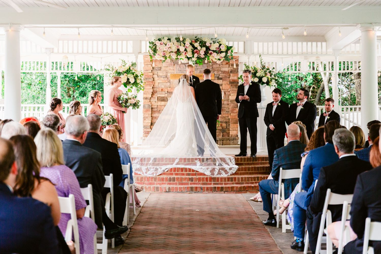 Wedding ceremony takes place under an elegant white porch with guests seated in rows facing the decorated altar.