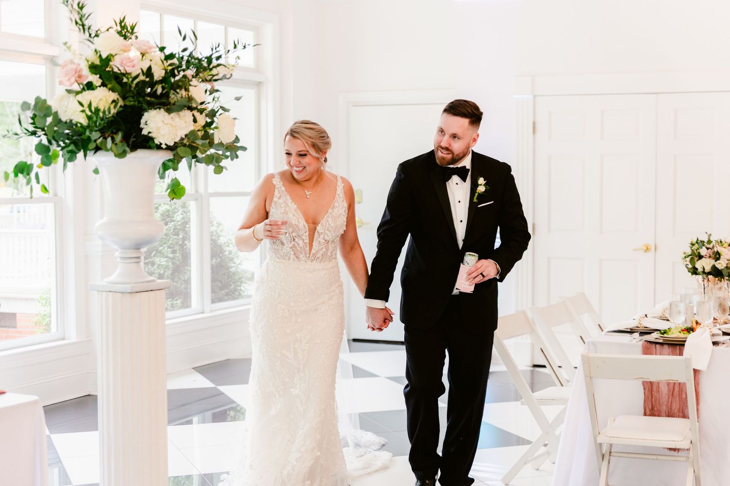 Elegant wedding reception table settings with white linens and floral centerpieces in a bright indoor venue.