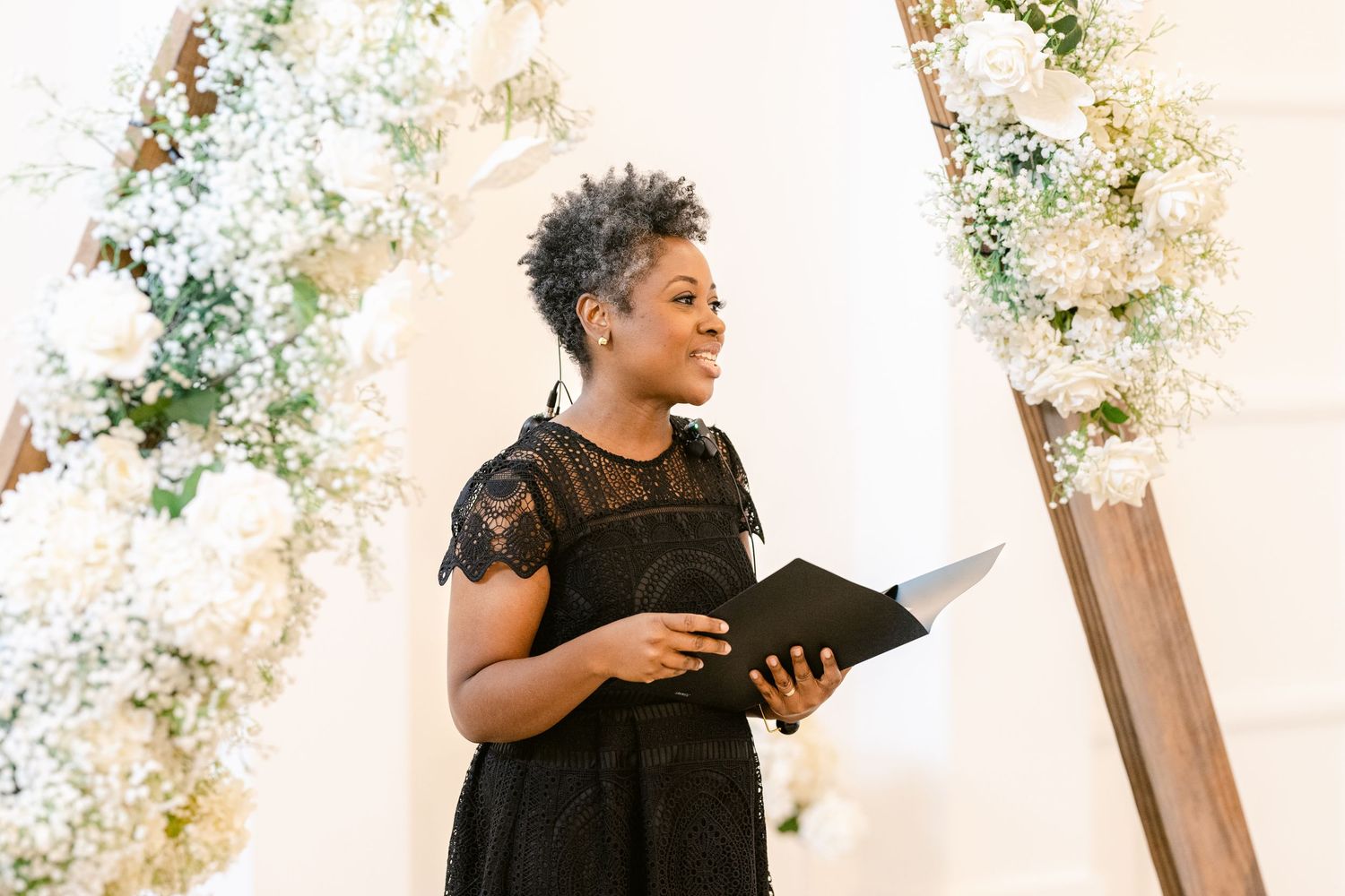 Wedding officiant in black lace dress reads from book at altar decorated with white floral arrangements.
