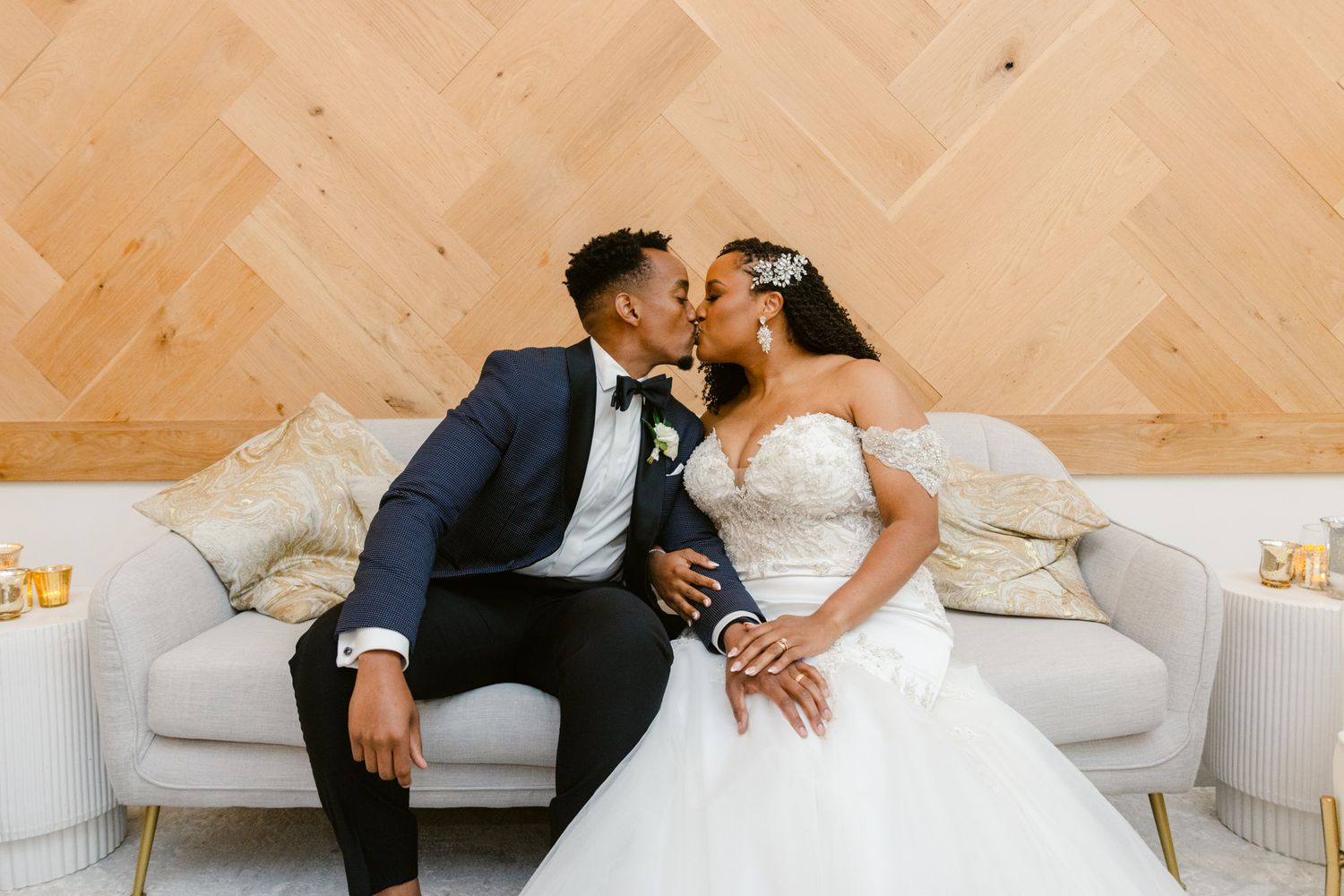 A romantic wedding moment with a couple sharing an intimate kiss while seated on a white couch against a chevron wood wall.