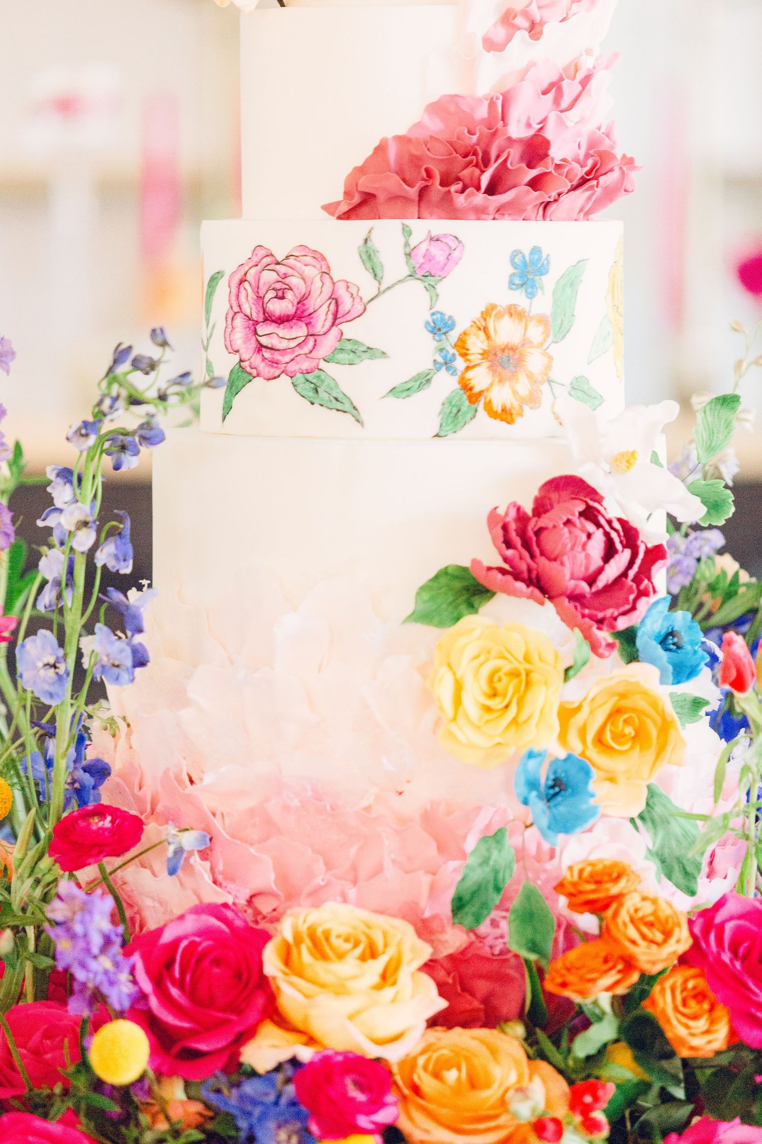 Colorful wedding cake decorated with hand-painted pink roses, yellow flowers, and blue delphinium.