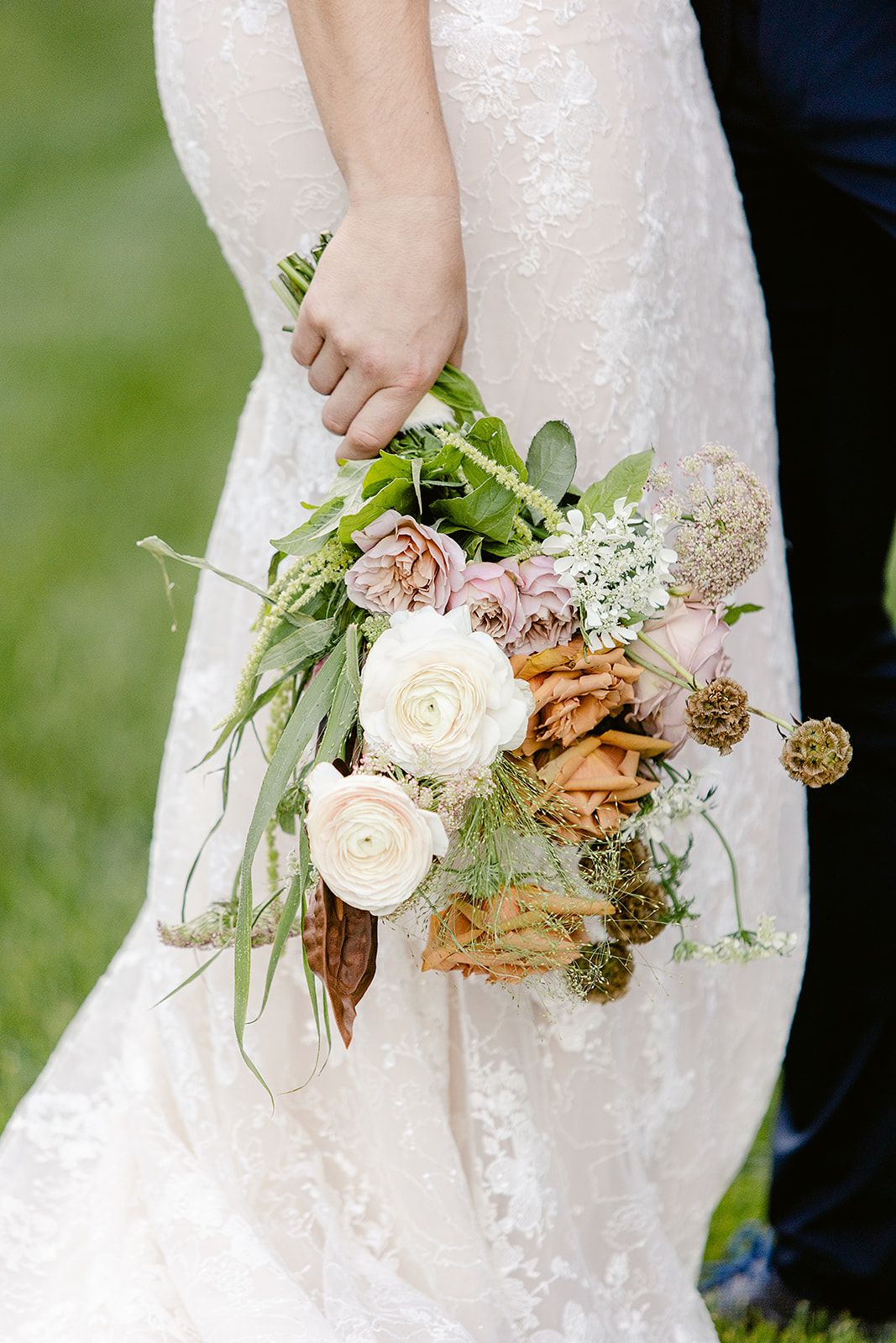 Elegant bridal bouquet with cream roses, copper-colored accents and sage green foliage against a white lace dress.