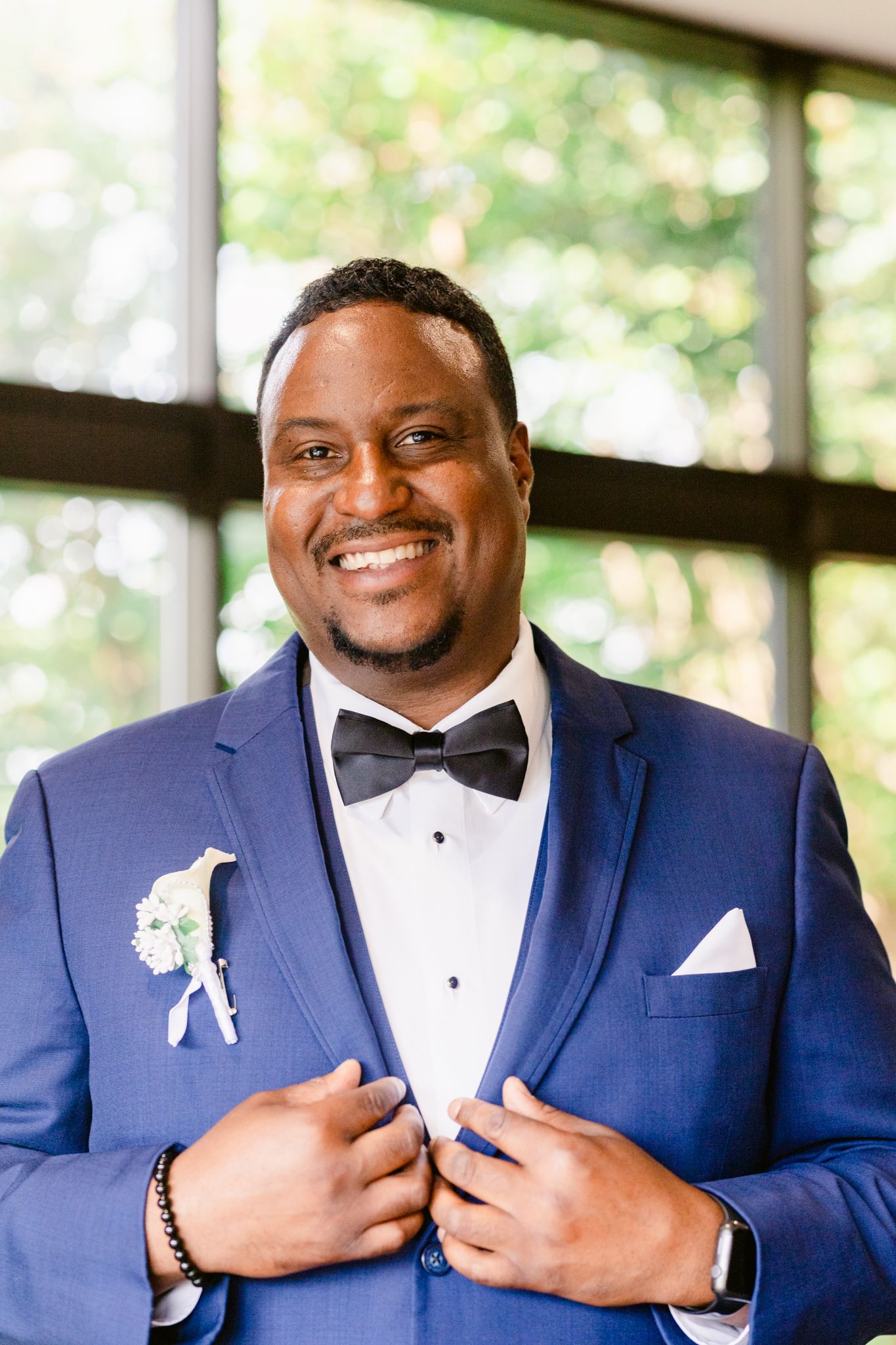 A person wearing a navy blue tuxedo with black bow tie and white pocket square smiles warmly in a bright indoor setting.