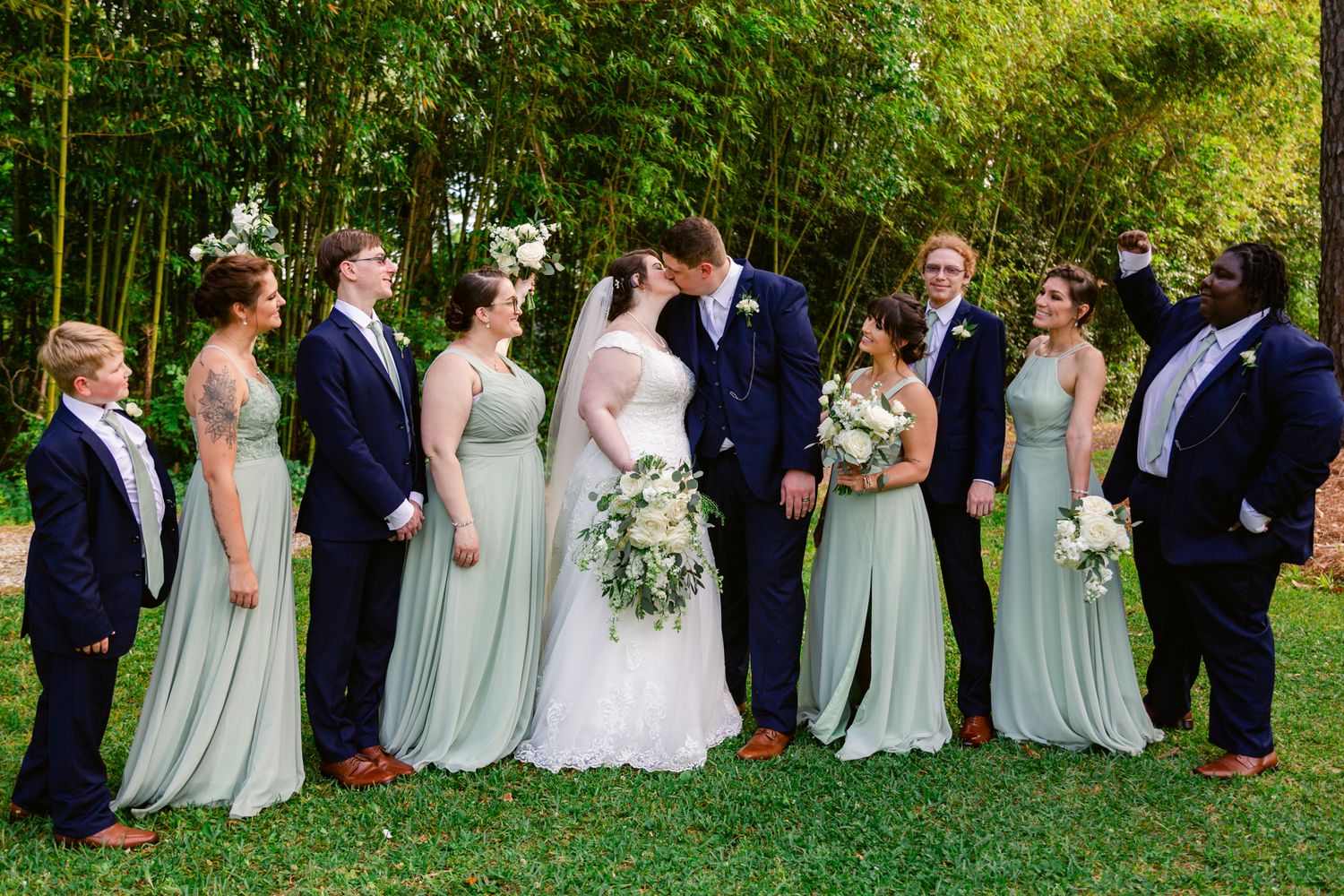 A wedding party in navy suits and sage green bridesmaid dresses poses outdoors in a line against lush greenery.