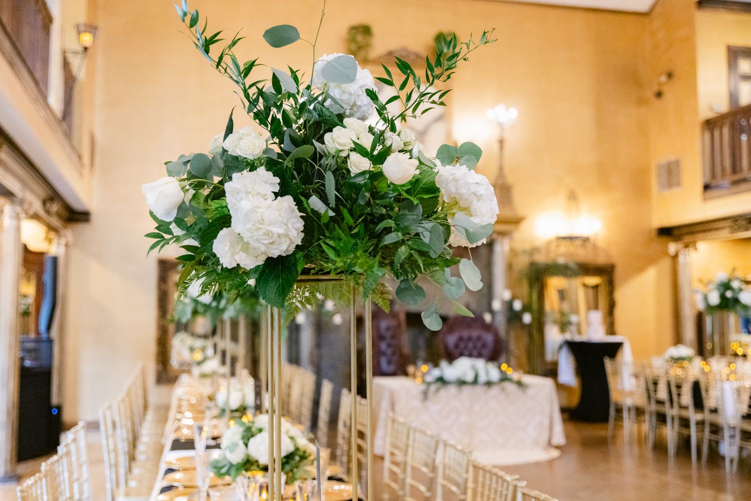 Elegant wedding reception hall decorated with white floral centerpieces and greenery in warm lighting.
