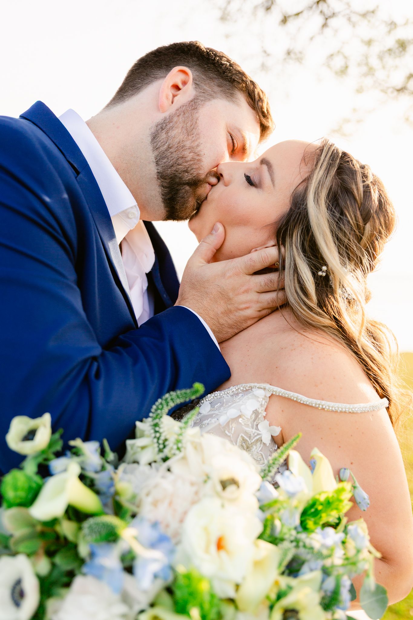 Romantic wedding couple sharing a tender kiss at sunset with white and blue floral bouquet in foreground.