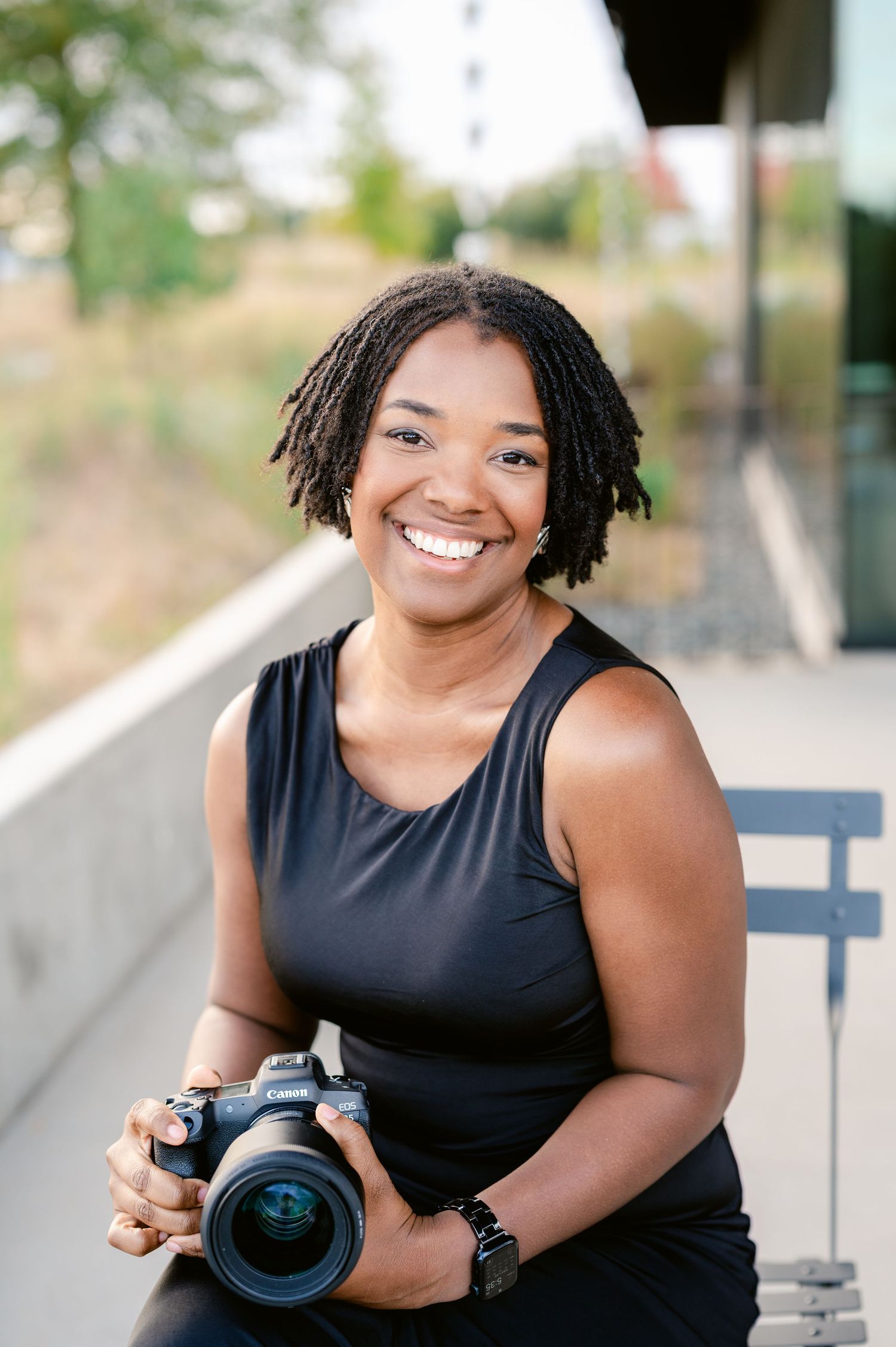 Professional photographer in a black sleeveless top holds a camera while smiling outdoors.