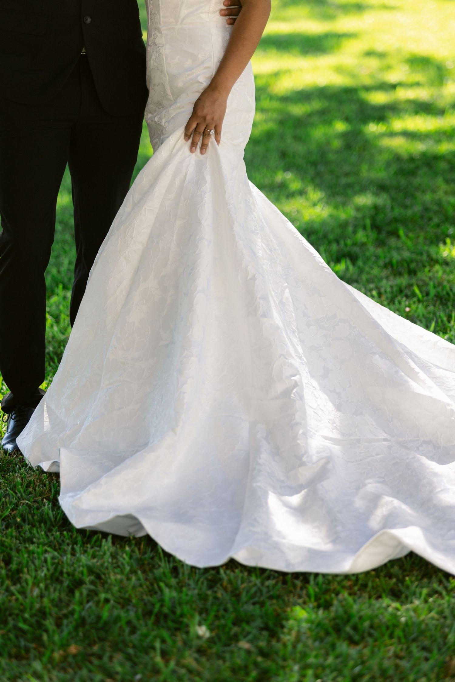 The bottom portion of a flowing white wedding dress twirls gracefully against lush green grass in a garden setting.