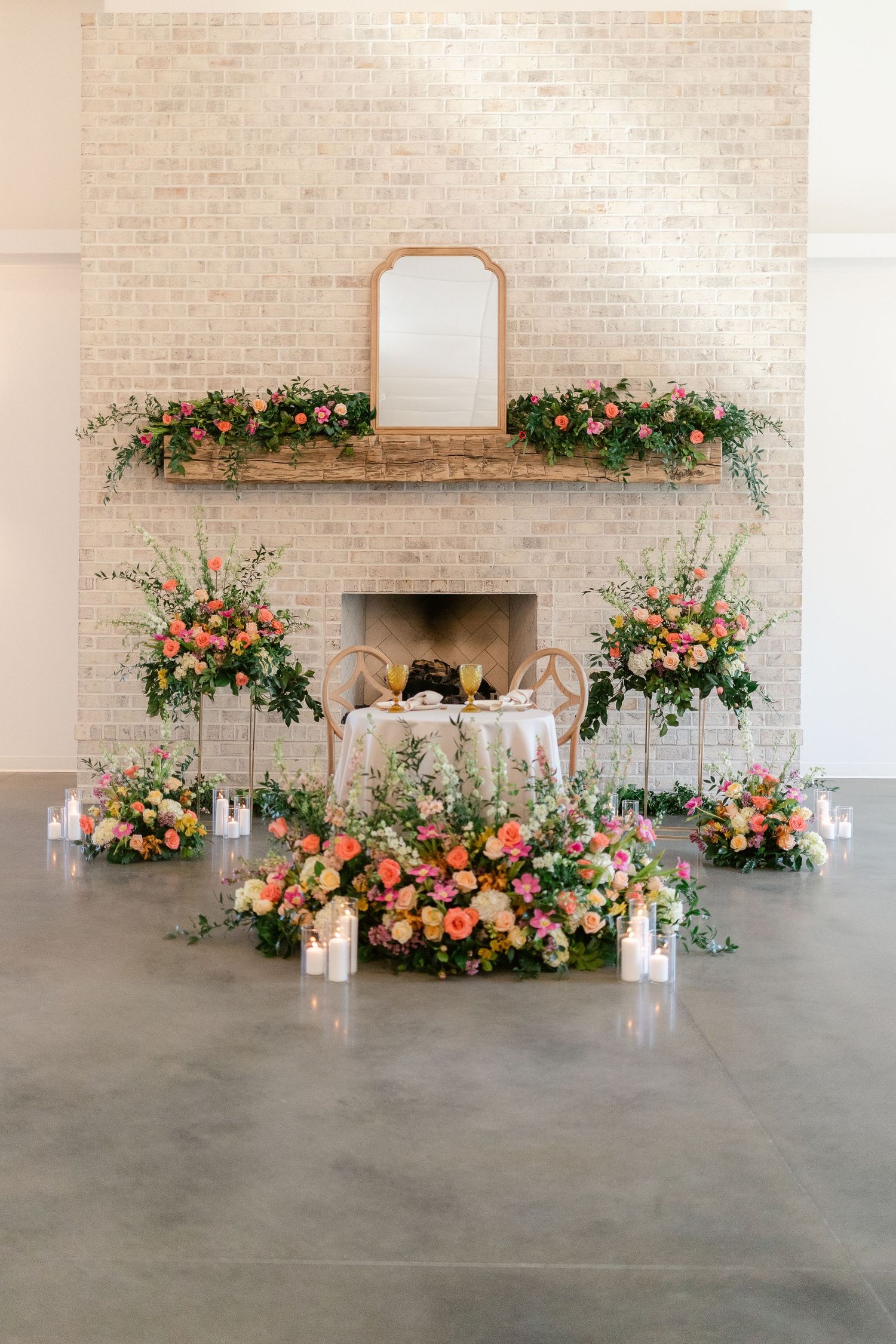 Elegant fireplace decorated with lush coral and peach floral arrangements and a mirror on white brick wall.