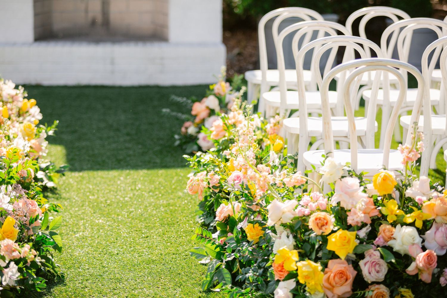 White chairs and colorful floral arrangements line a grassy outdoor wedding ceremony space in elegant garden setting.