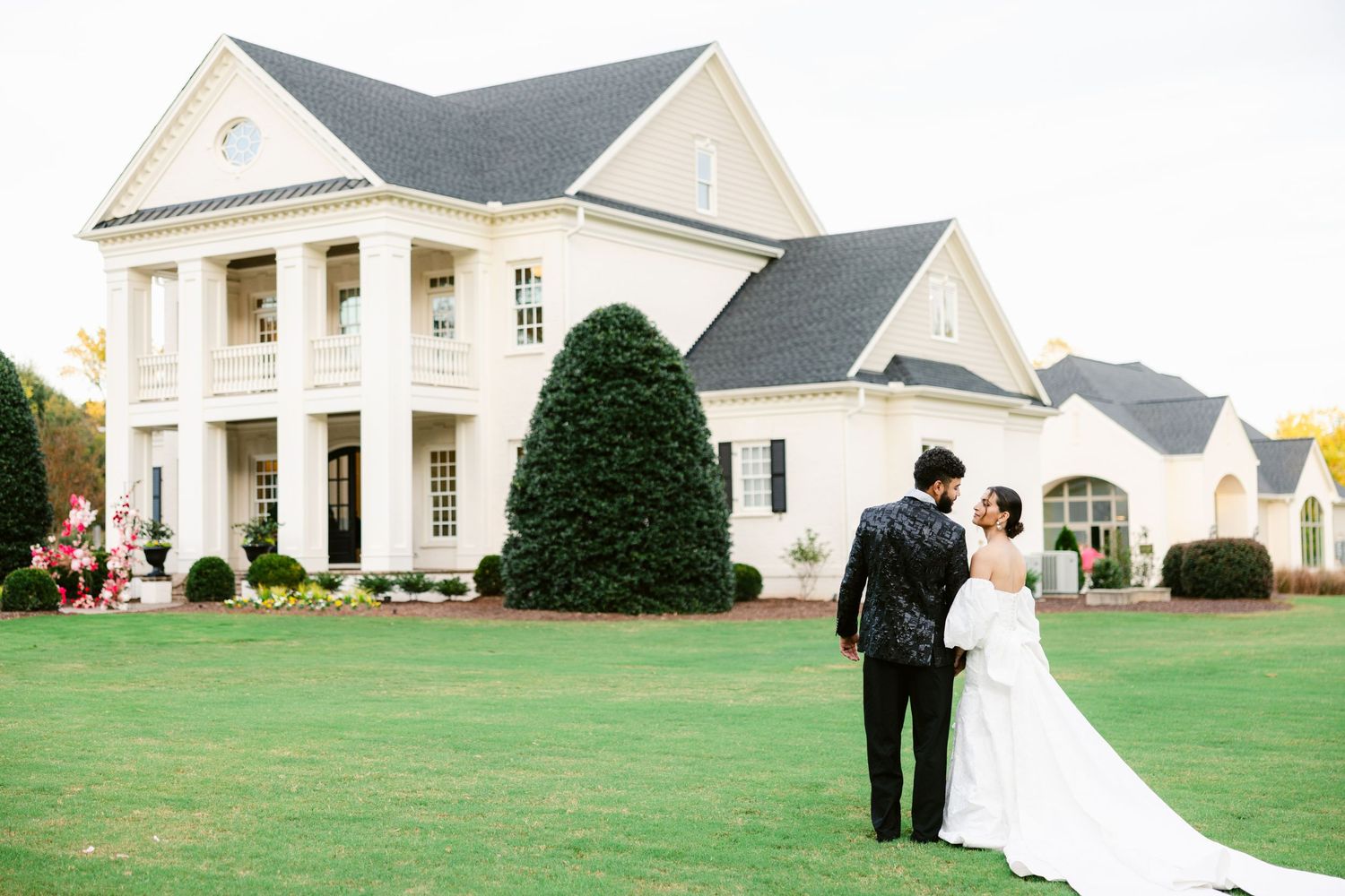 Wedding couple walks across a manicured lawn in front of an elegant white colonial style mansion.
