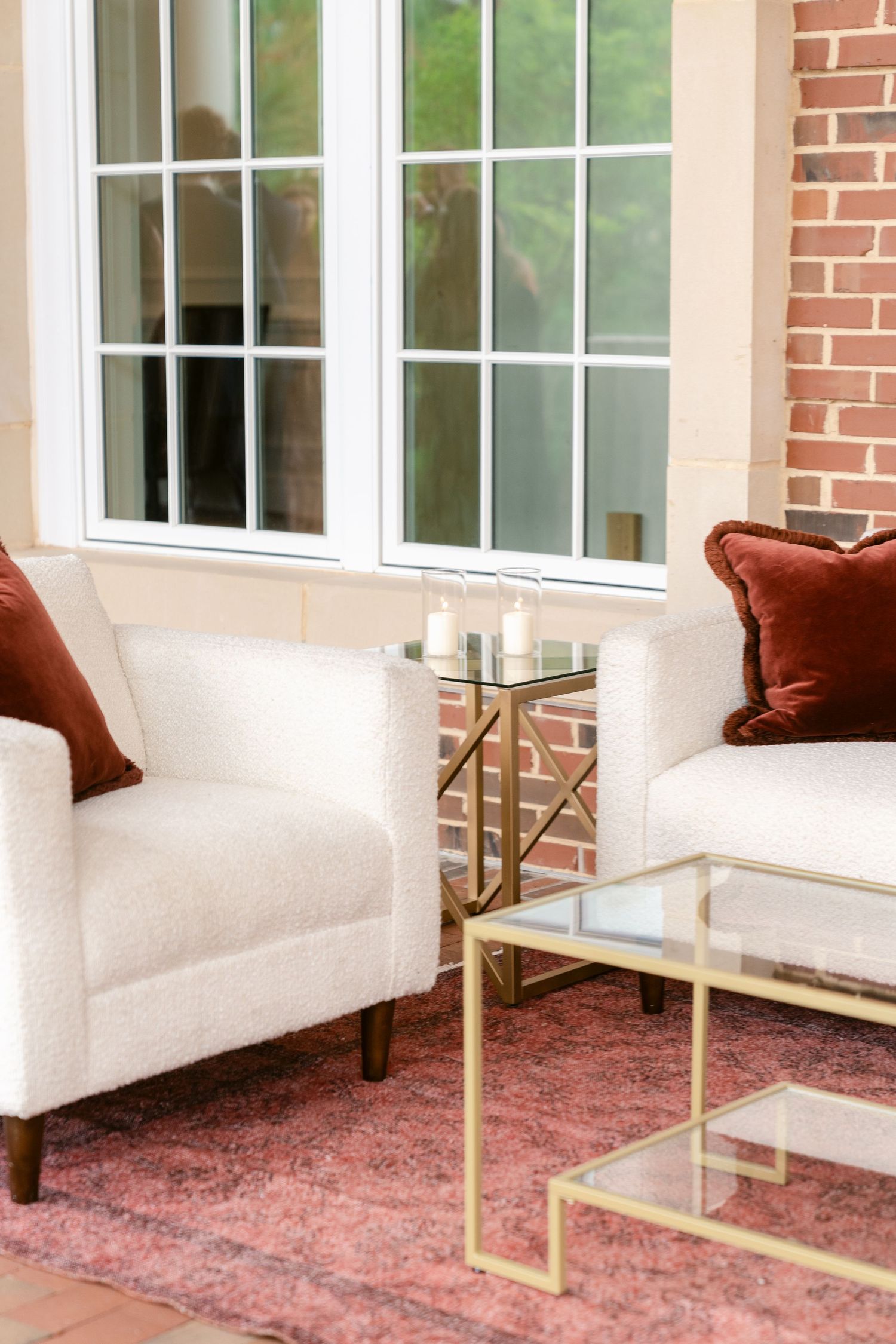 Modern white upholstered bench with gold metal frame and burgundy throw pillows sits against brick wall by windows.