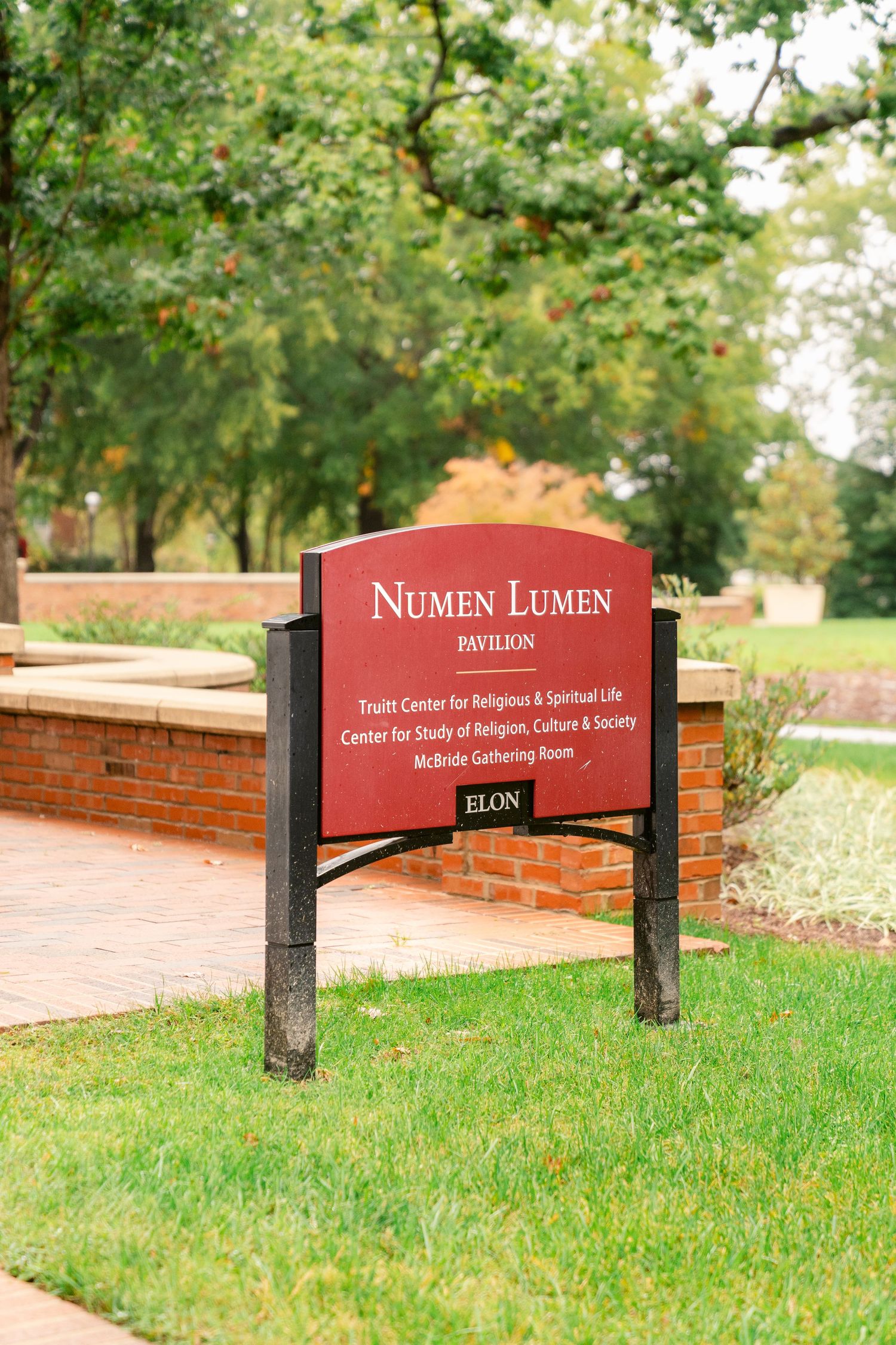 A red Numen Lumen campus sign stands in front of a green lawn and trees at Elon University.