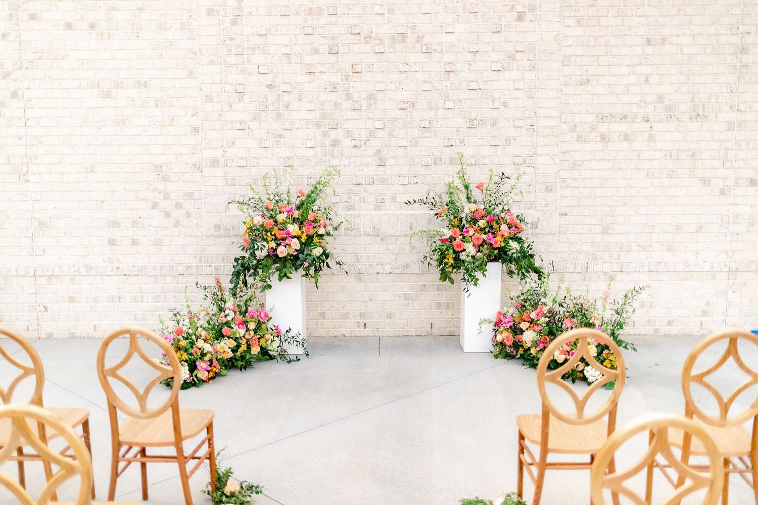 Modern white wedding ceremony setup with geometric wooden chairs and floral arrangements in a bright indoor venue.