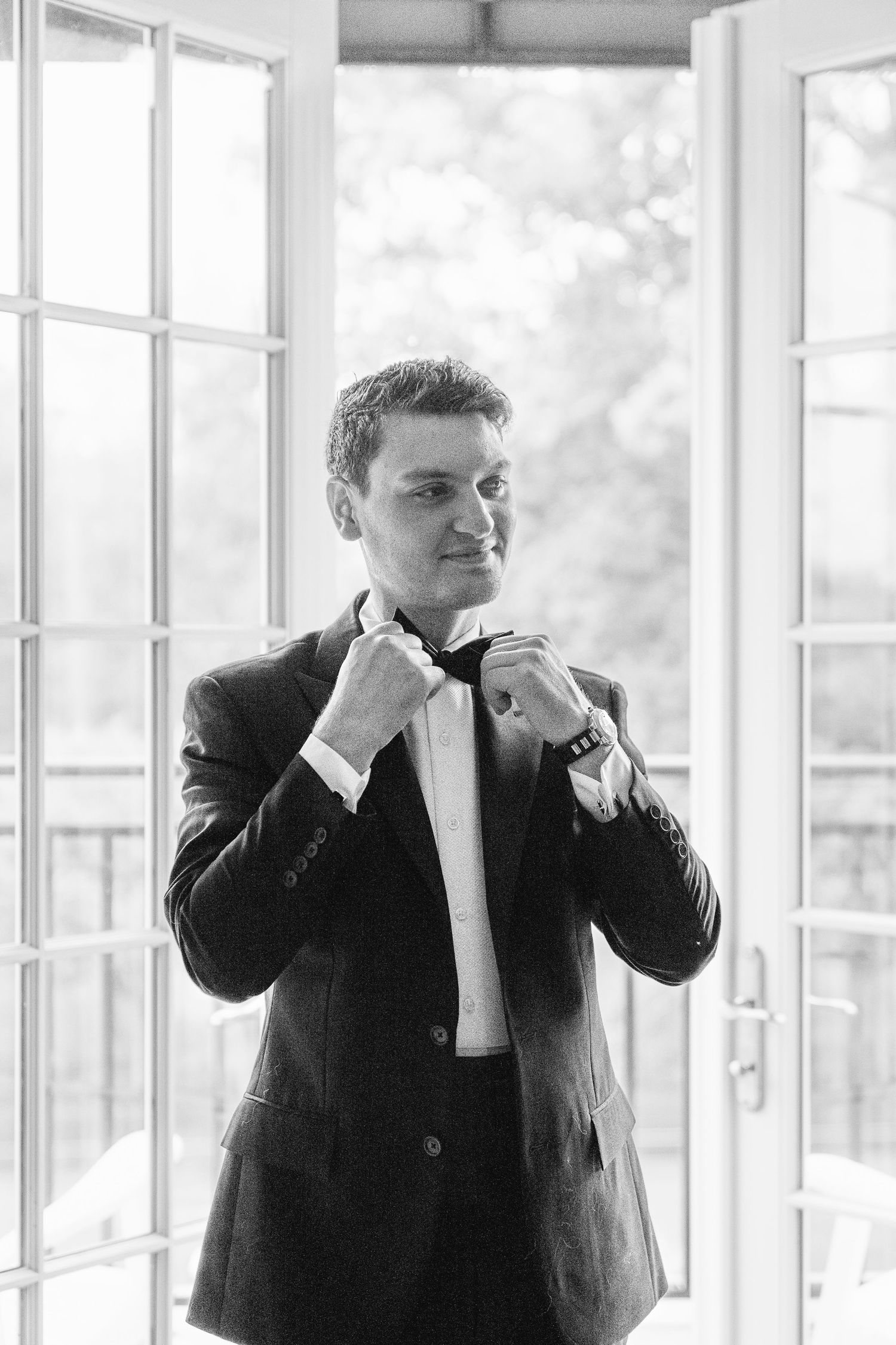 A person in a black suit and white shirt stands in a bright room with large windows, making expressive gestures.