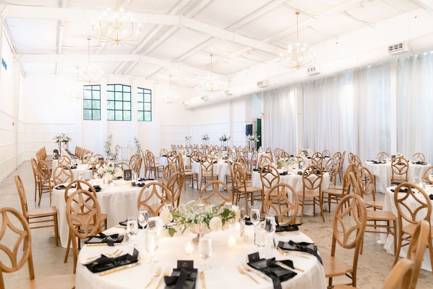 Elegant wedding reception venue with white decor, wooden chairs, and round tables set for a formal celebration.