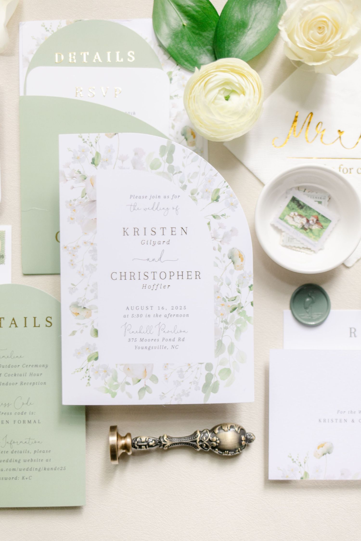 Elegant wedding invitation suite in white and sage green with floral details and gold accents laid out on cream surface.