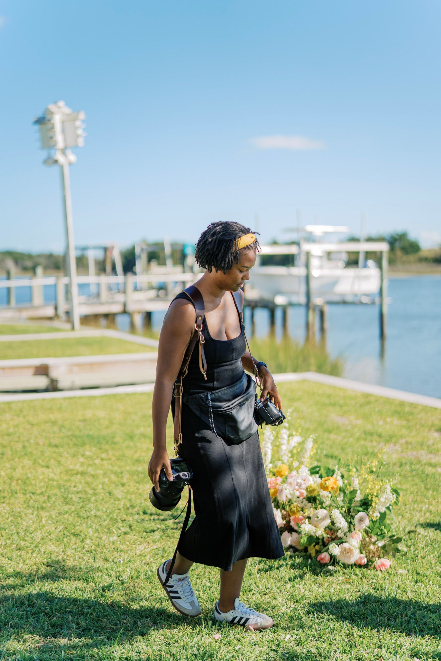 A figure in a black dress kneels on grassy waterfront with a colorful bouquet of flowers beside them.
