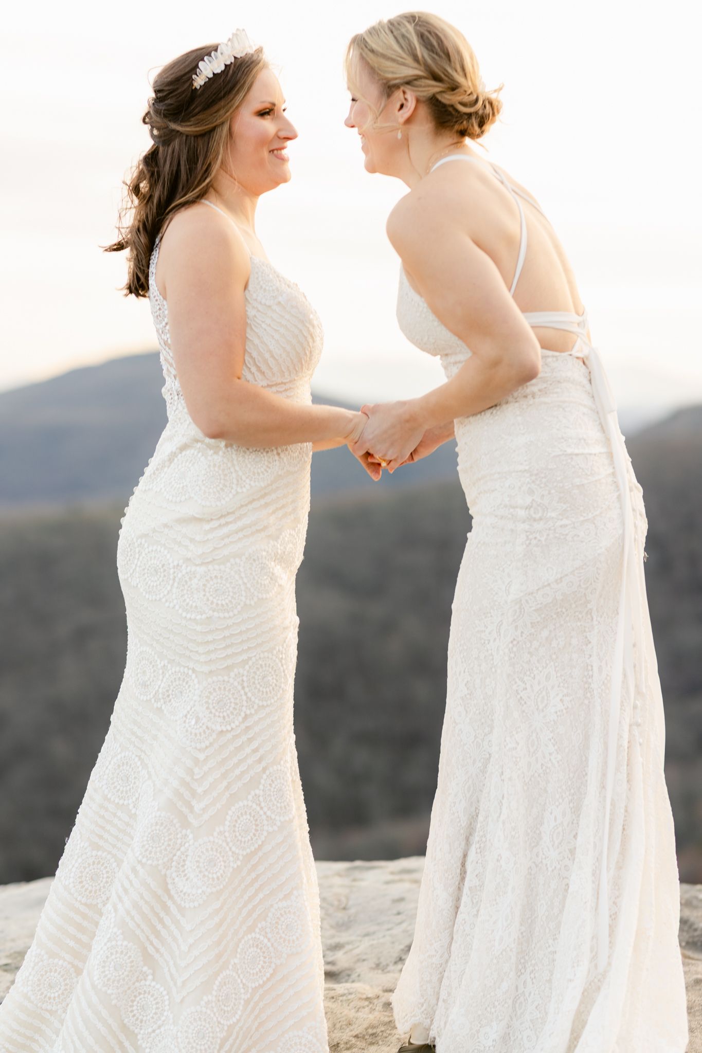 Two people in white wedding dresses share a joyful moment together on a mountaintop overlook.