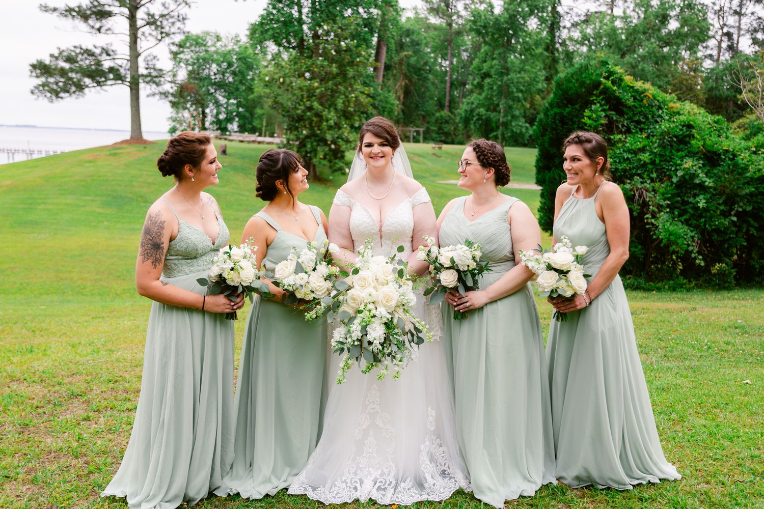 Bridesmaids in sage green dresses stand with bride in white gown against lush outdoor backdrop.