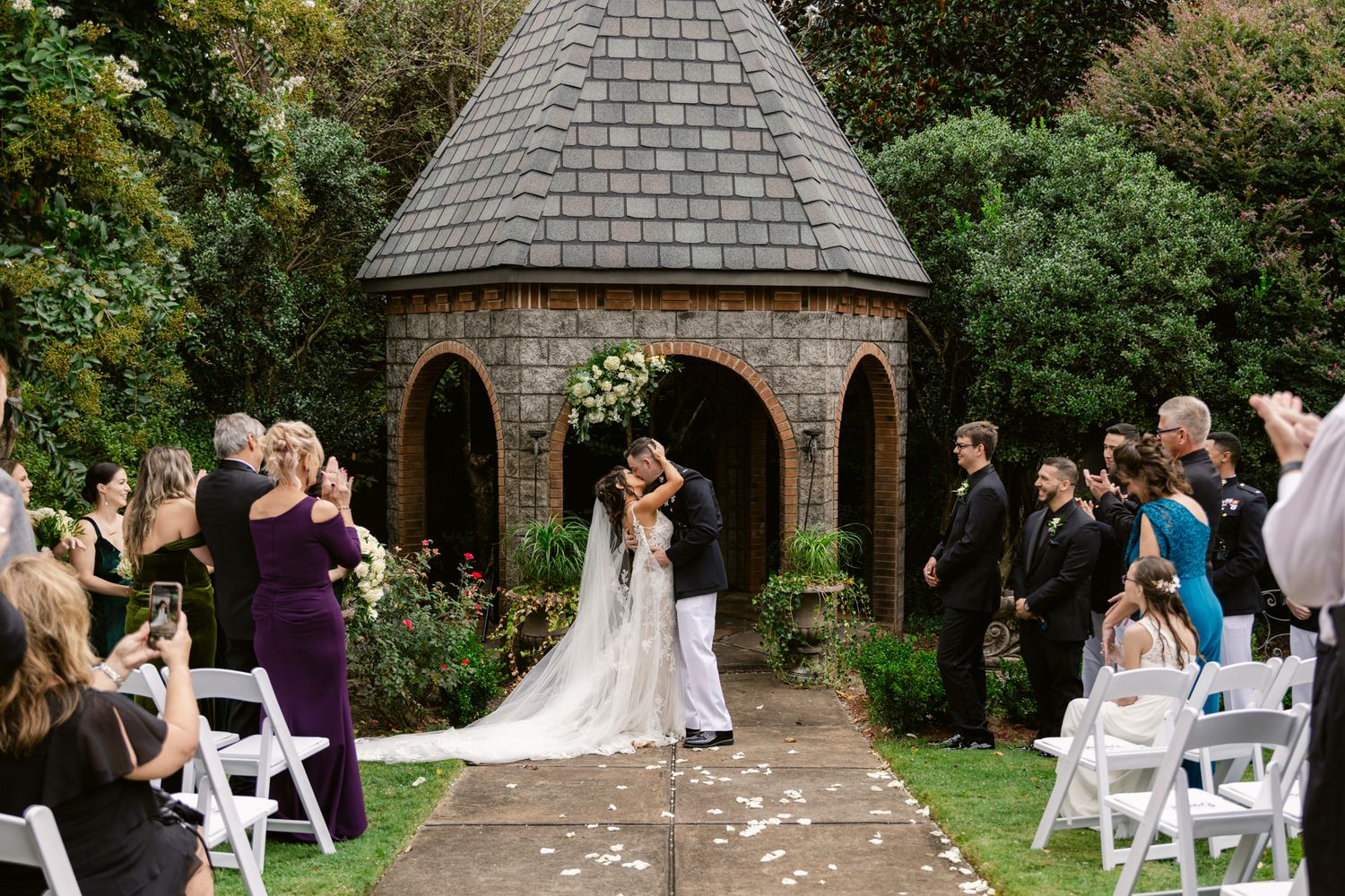 A wedding ceremony setup with white chairs in a garden courtyard featuring a rustic stone gazebo and archway.