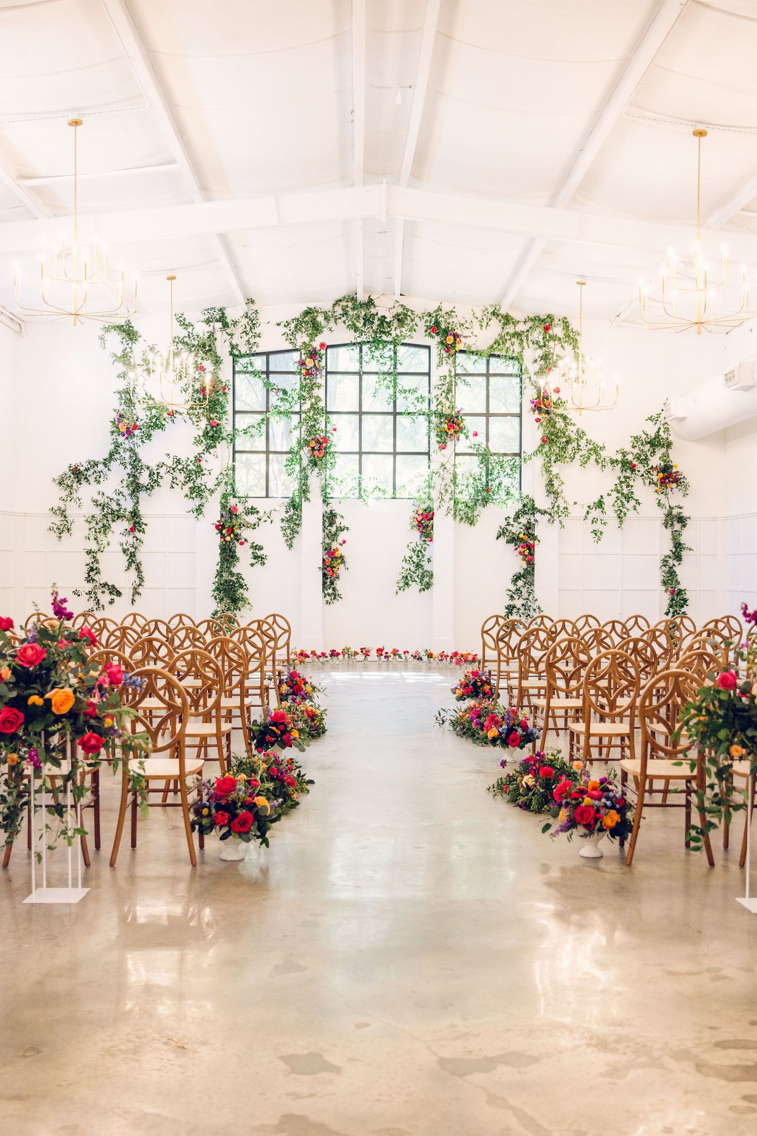Indoor wedding ceremony setup with wooden chairs, pink floral arrangements, and greenery hanging from a window backdrop.