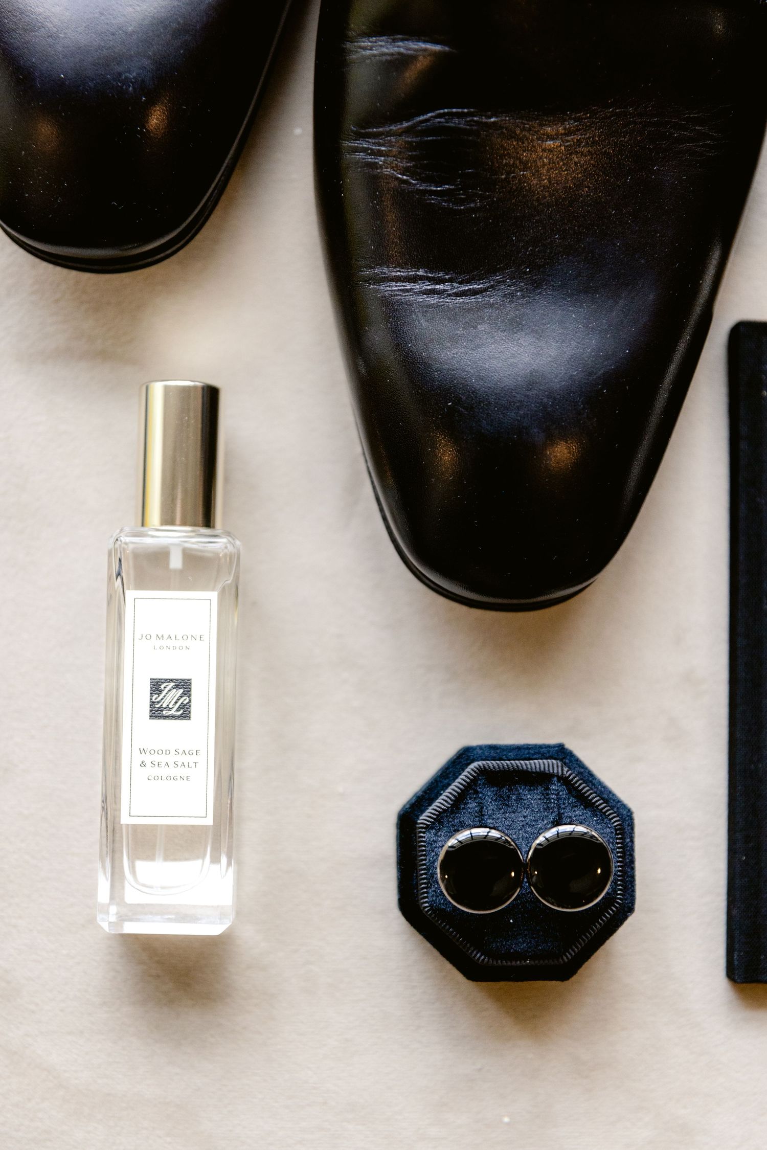 Wedding accessories including black dress shoes, rings, and perfume bottles arranged on white surface.