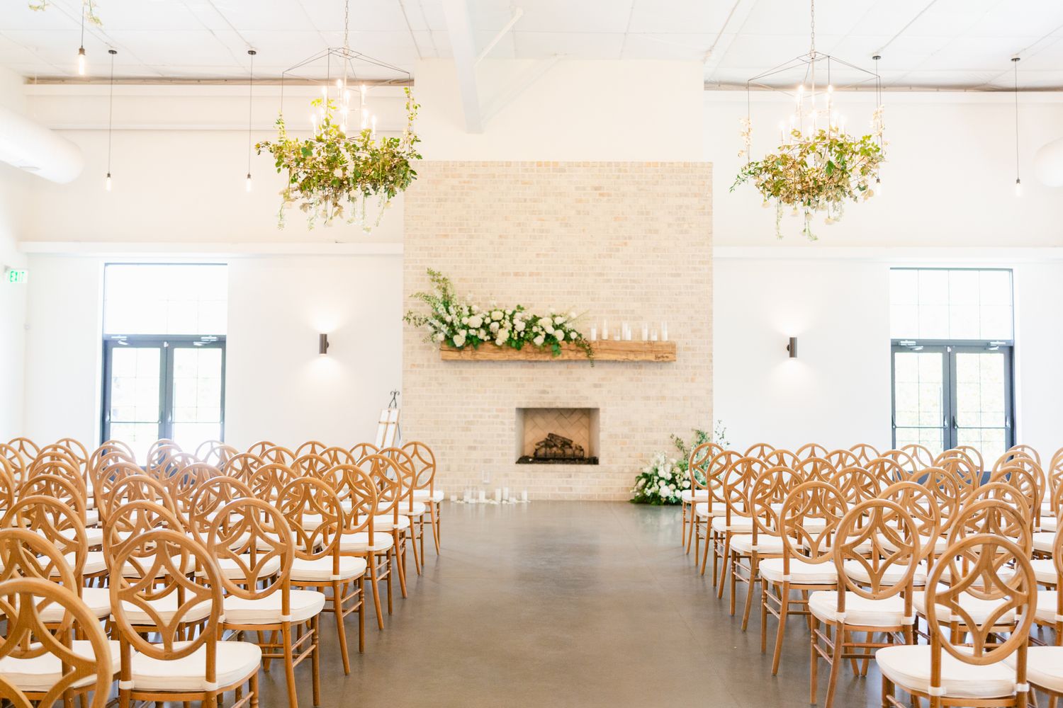 Elegant white wedding ceremony venue with wooden chairs and hanging floral arrangements in a bright modern space.