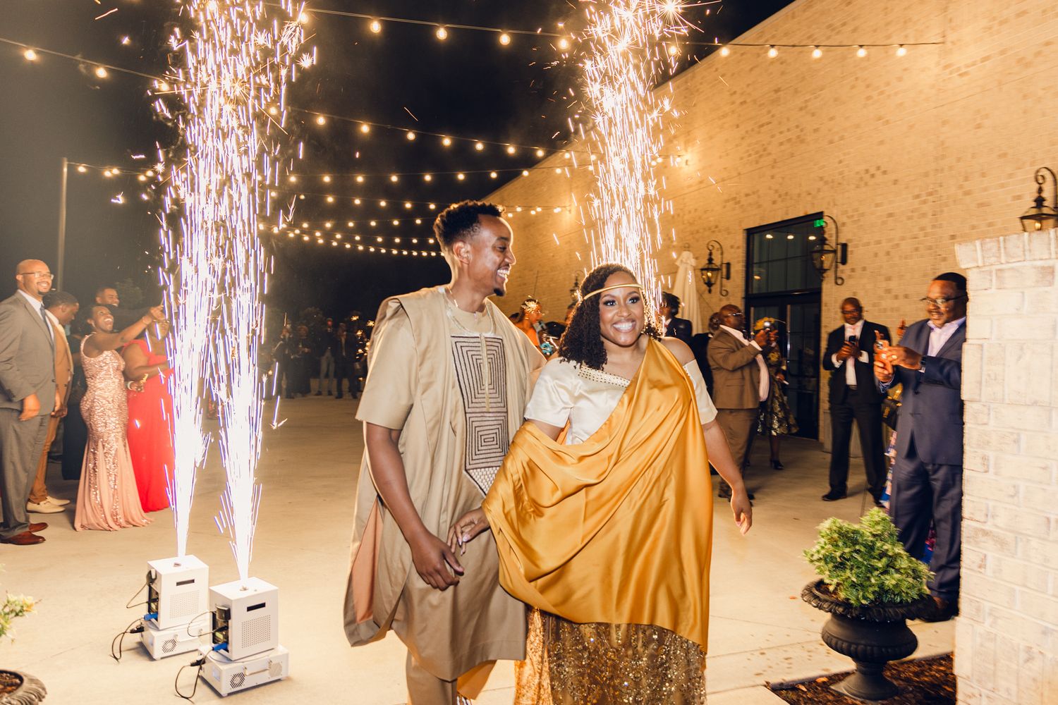 String lights and sparklers illuminate an evening wedding celebration against a light colored brick wall.