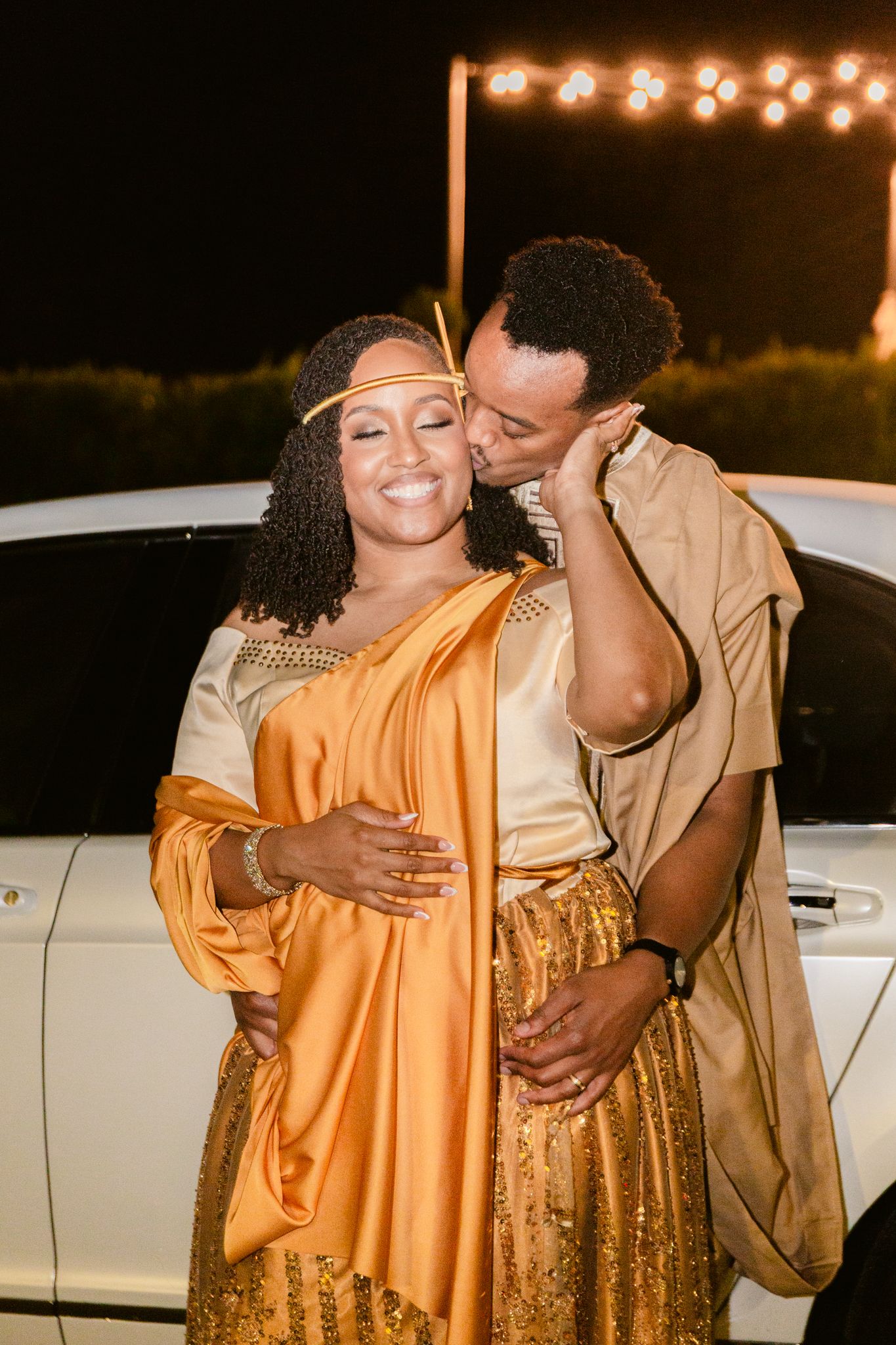 A couple in formal attire embrace and laugh together at an outdoor evening event with string lights visible.