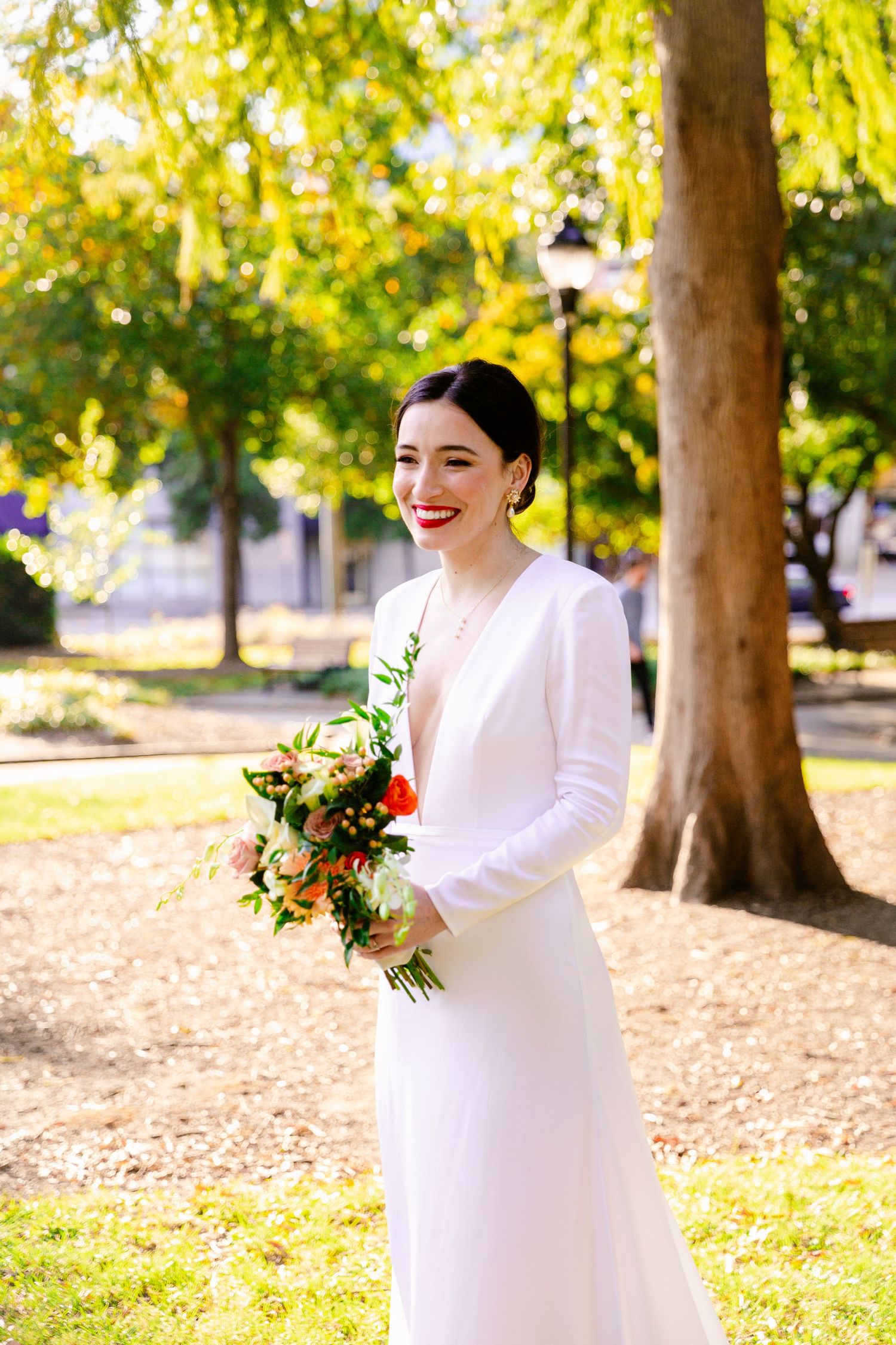 A bride in a long-sleeved white dress smiles while holding a bouquet in a sunlit autumn park with golden leaves.