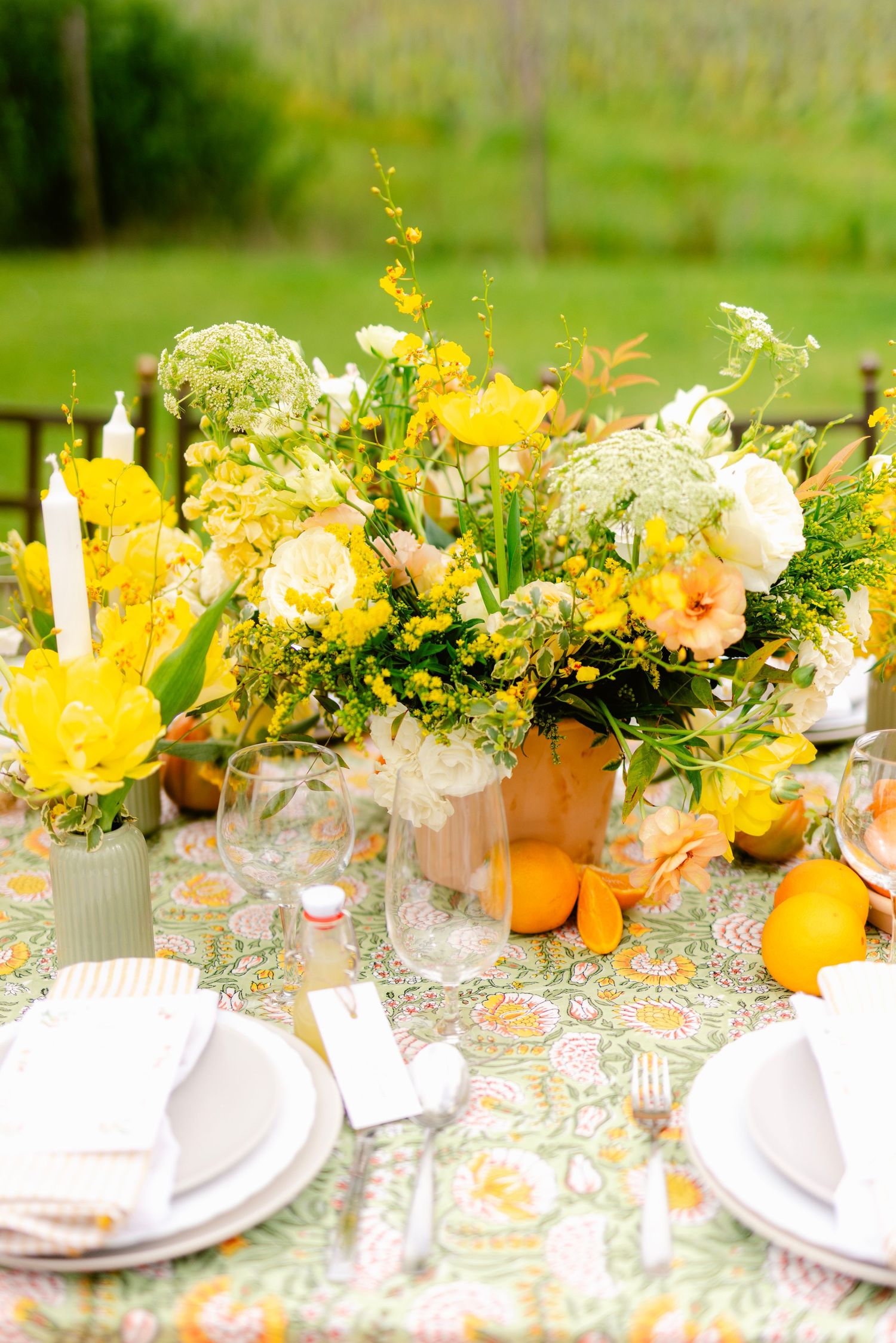 An elegant outdoor table setting with yellow and white floral arrangements, pumpkins, and place settings on a floral tablecloth.