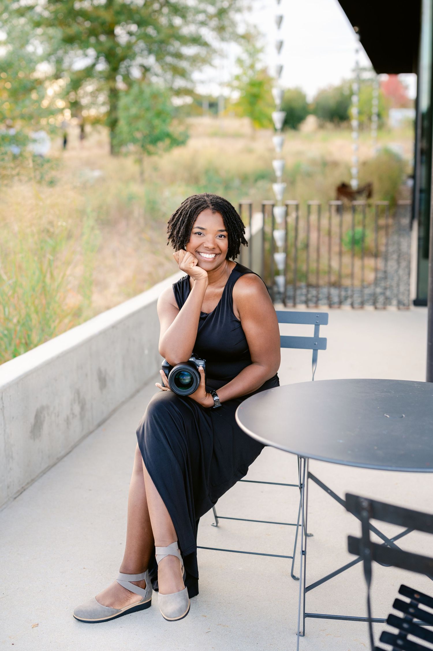 A person in a black dress sits on outdoor furniture while taking photos with a professional camera in a sunny setting.