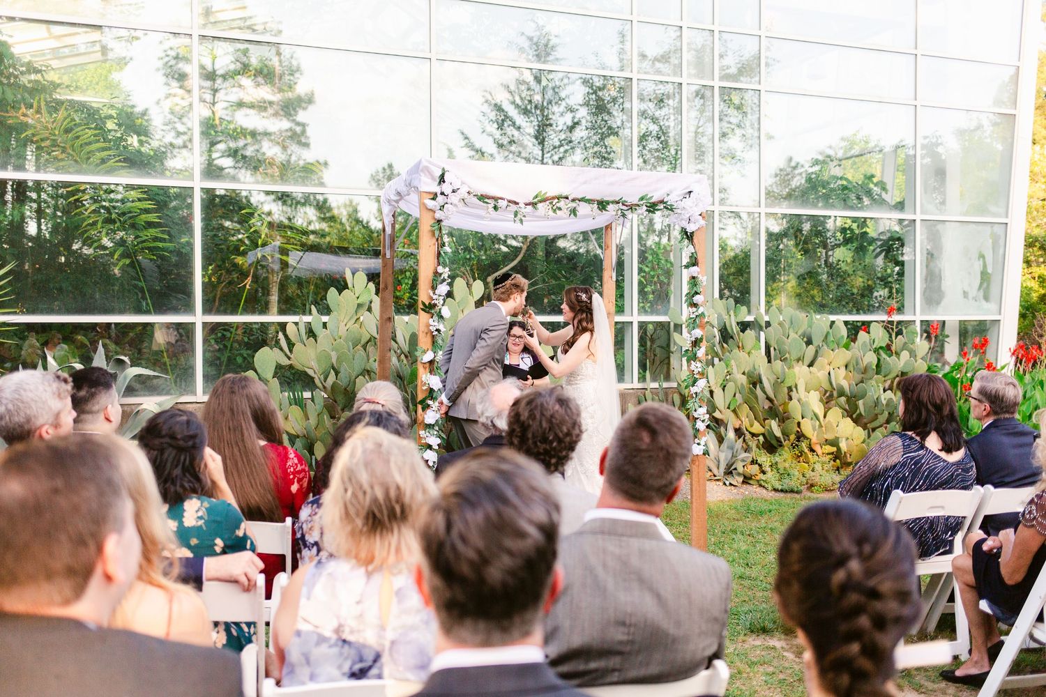Outdoor wedding ceremony taking place in a glass conservatory with lush greenery visible through the windows.
