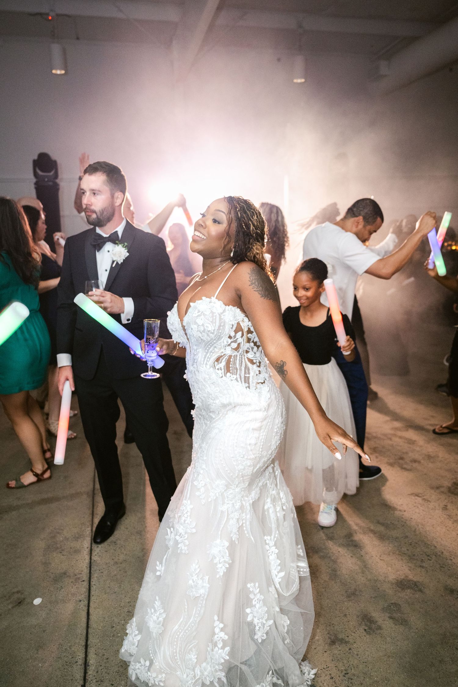 A person in a white lace wedding dress dances joyfully at a reception celebration with other guests on the dance floor.