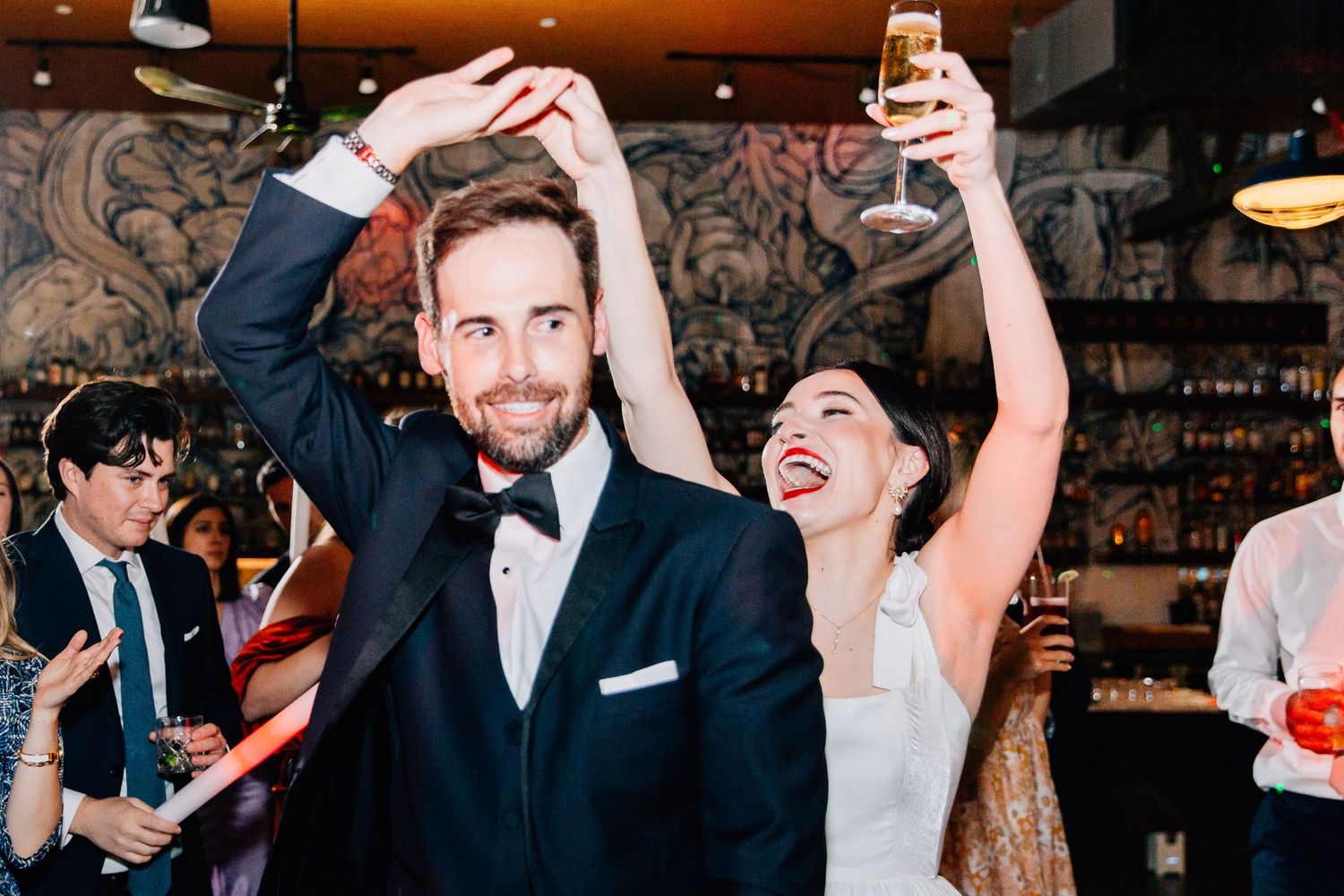 Joyful bride and groom dancing and raising glasses at elegant wedding reception in stylish bar venue.