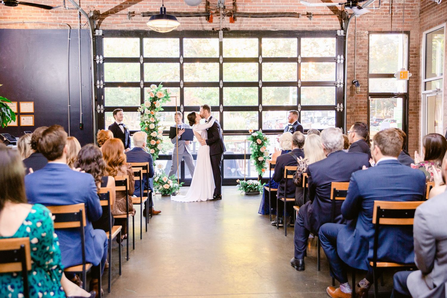 People sitting in rows at tables in a large conference or dining room with exposed brick walls and industrial lighting.