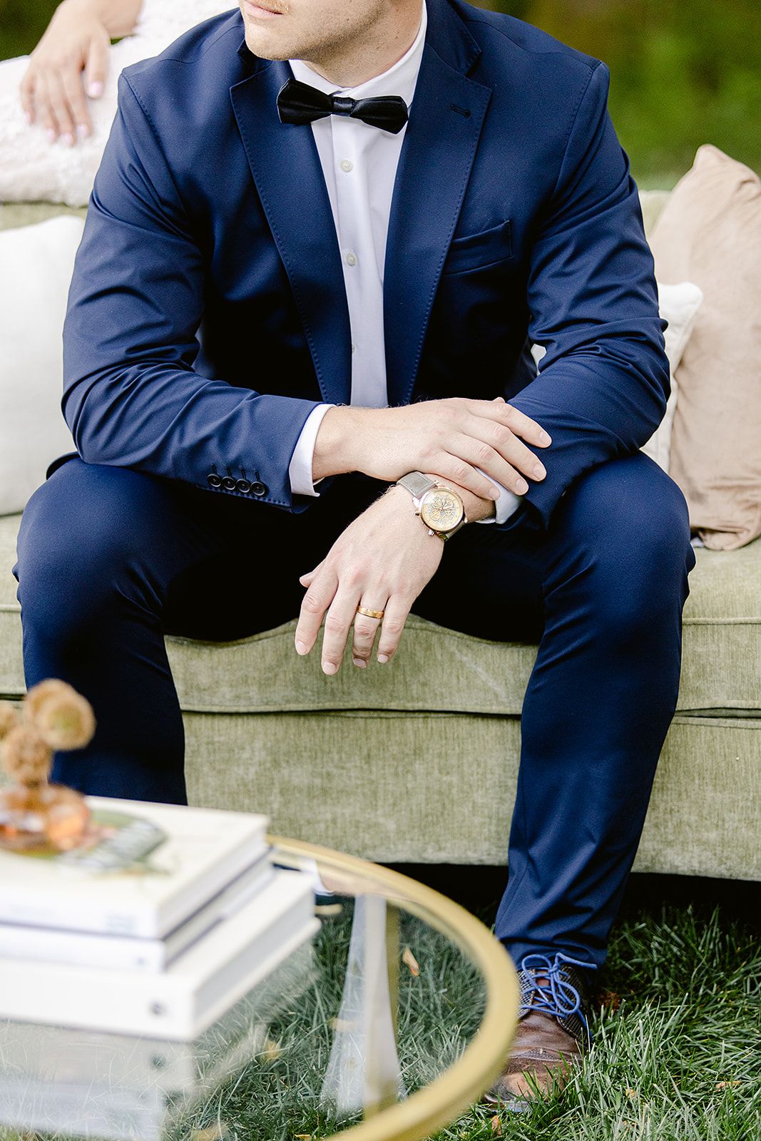 Person in navy blue suit and bowtie sitting on outdoor bench with polished dress shoes.