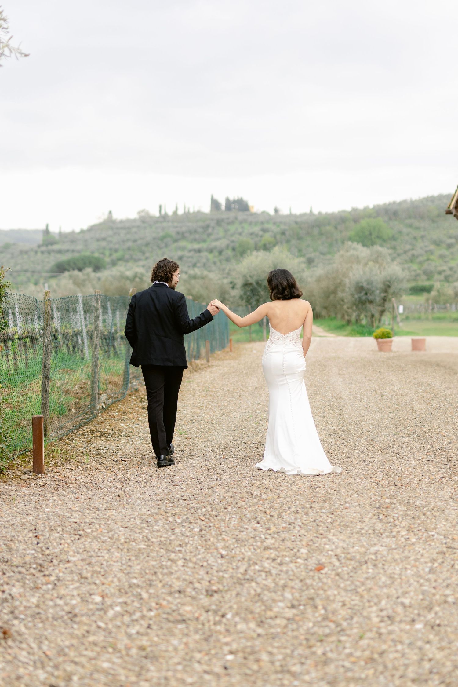 A couple walks hand in hand down a gravel path at a scenic vineyard estate.
