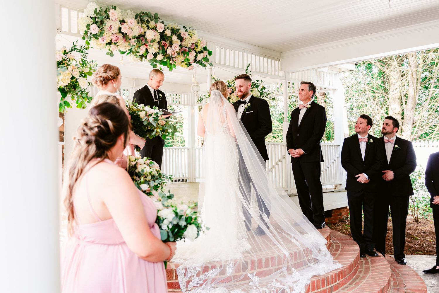 Wedding ceremony under a white covered porch with floral arch and pink bridesmaid dresses.