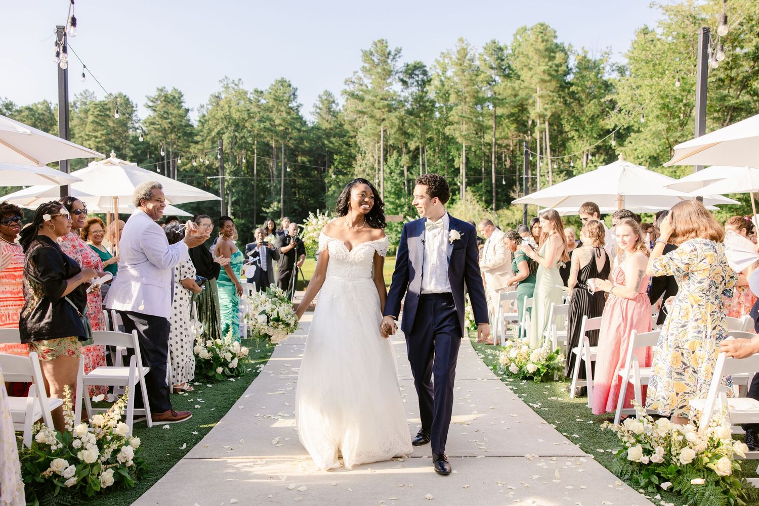 Panoramic view of an outdoor wedding reception with guests mingling on a lawn surrounded by trees and white tents.