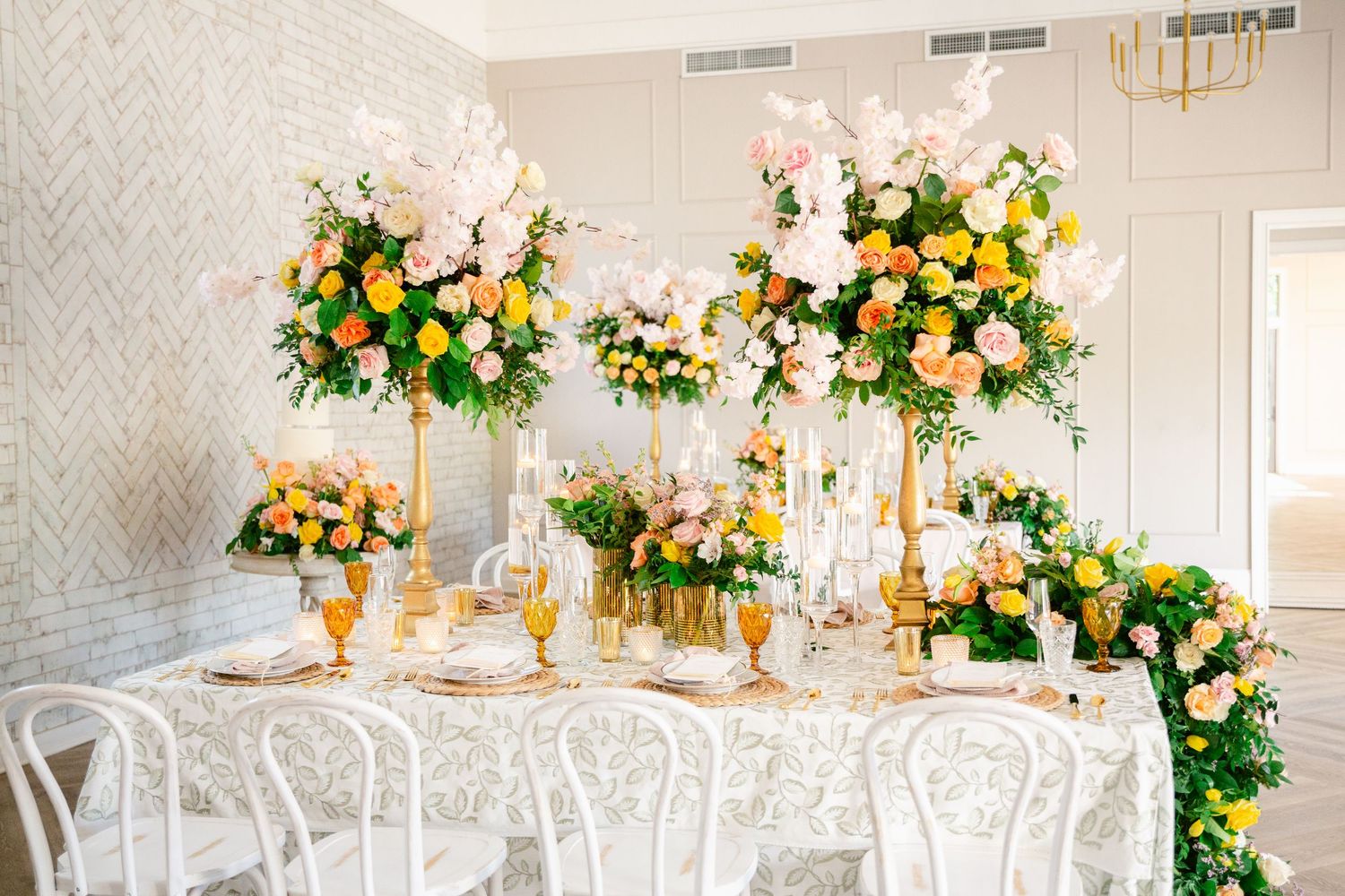 Elegant white wedding reception tables with floral centerpieces featuring pink and peach roses in a light-filled ballroom.