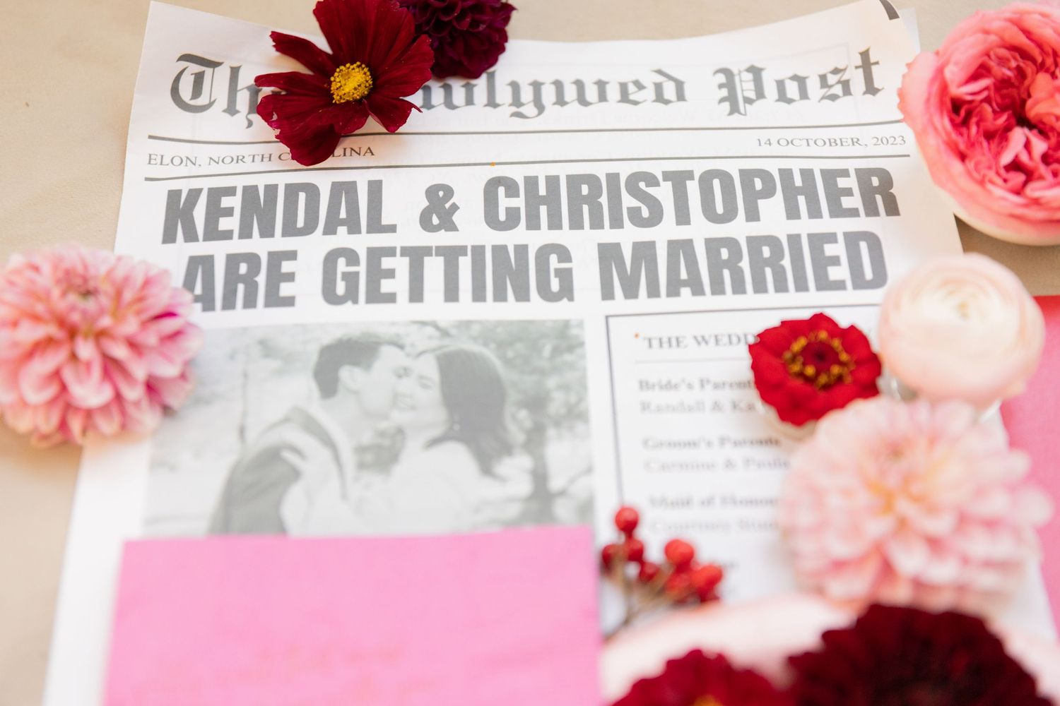 Wedding invitation details surrounded by pink and red flowers including roses and carnations on a light background.