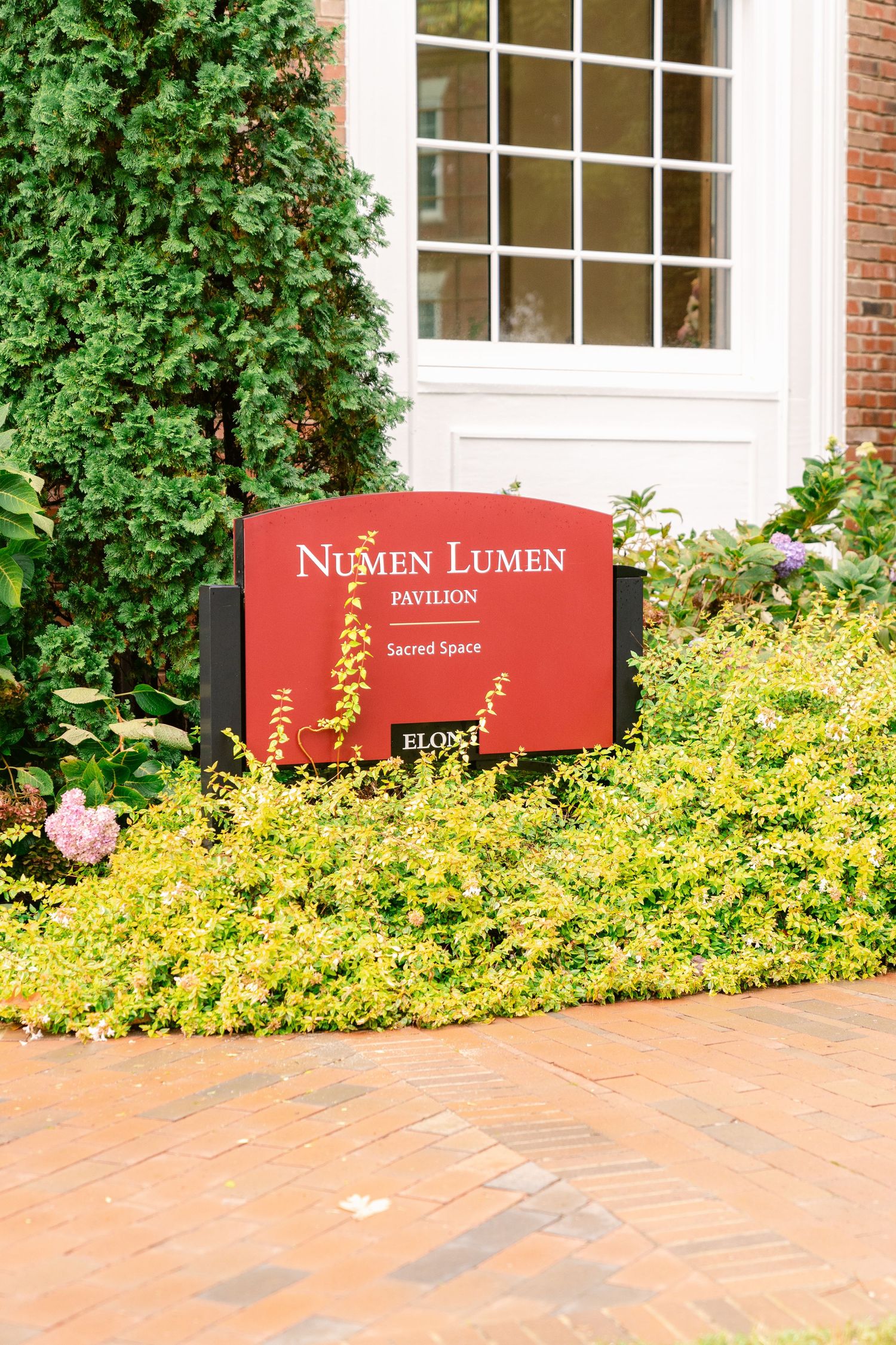 Red Numen Lumen pavilion sign surrounded by yellow flowers and green foliage on a brick pathway.