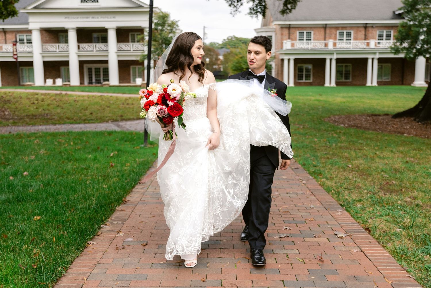 Newlyweds walk together along a brick pathway through a lush green lawn on their wedding day.