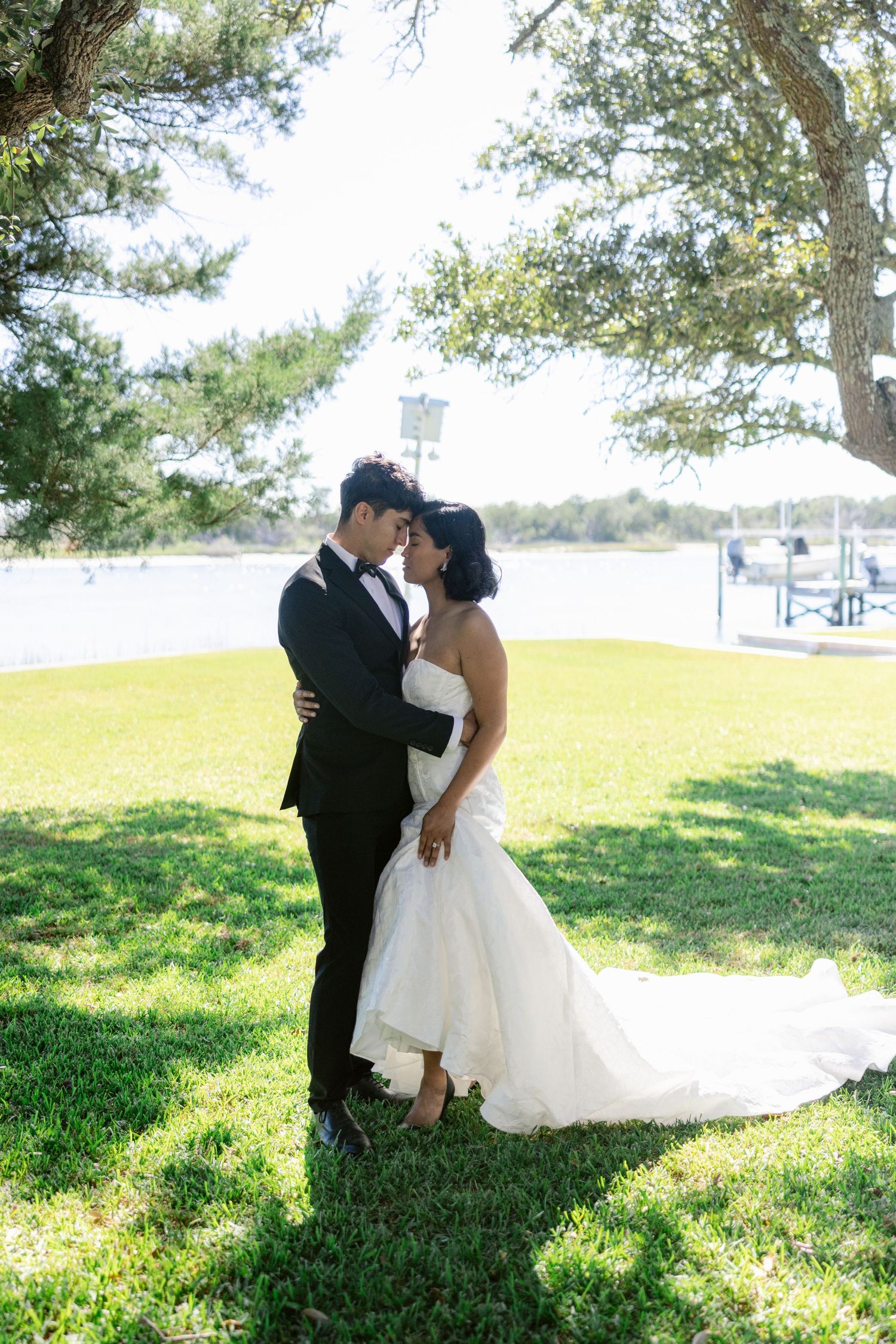 A bride and groom share romantic moments on a sunny hillside with green grass and trees in the background.