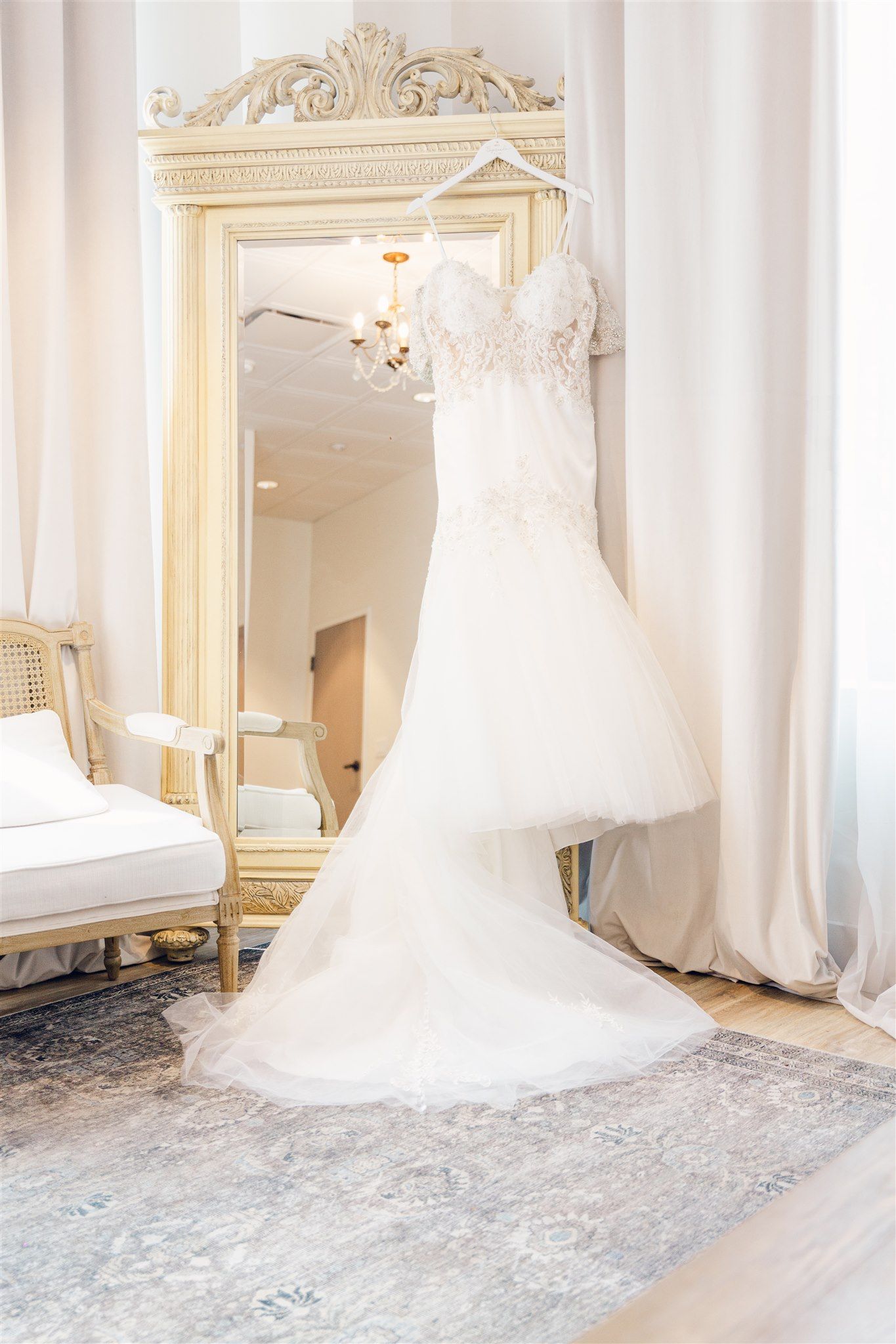 White wedding dress with long train displayed in an elegant room with ornate gold mirrors and white decor.